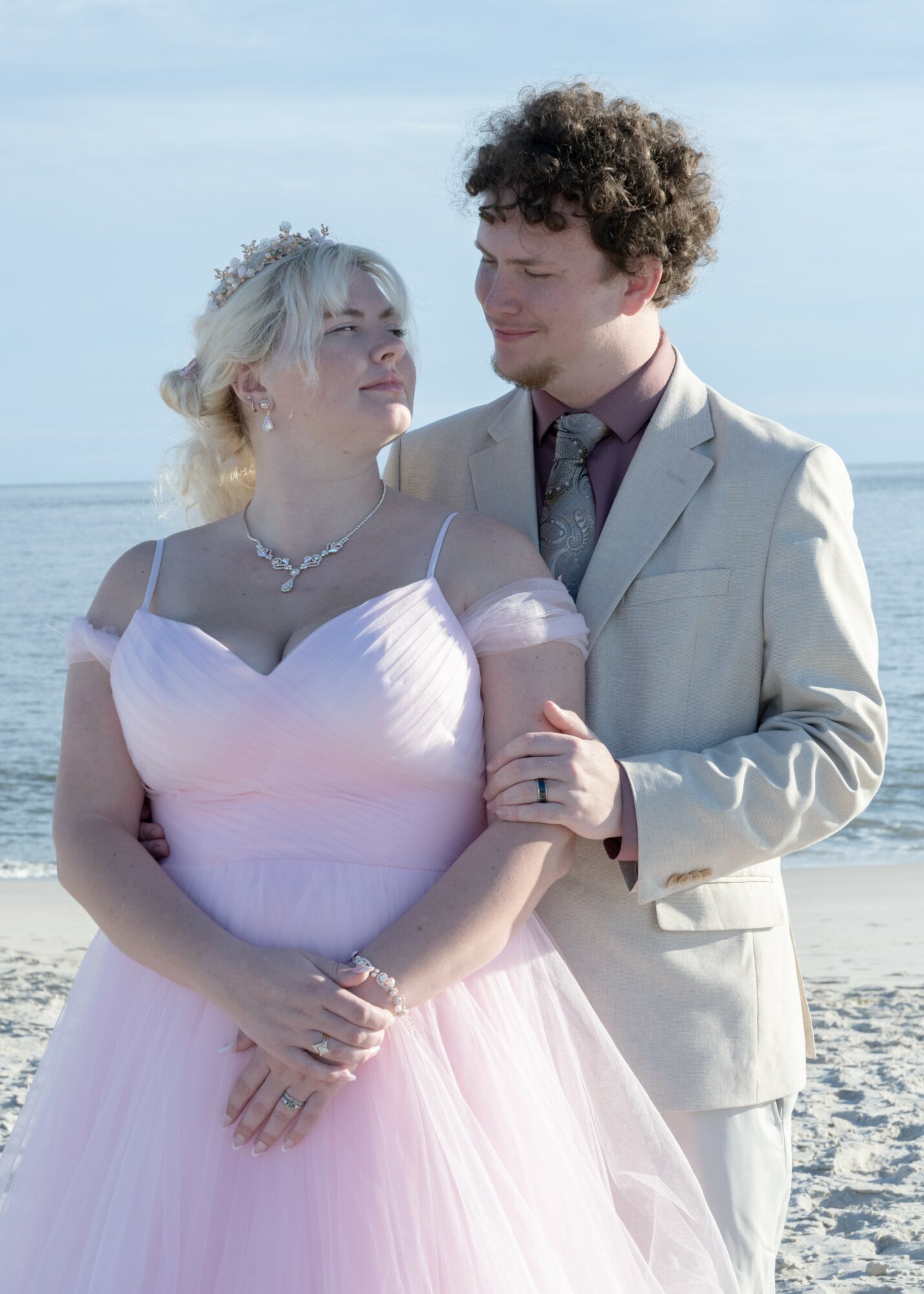 A woman in a pink dress and a man in a light suit stand close on a beach, holding hands and looking at each other.