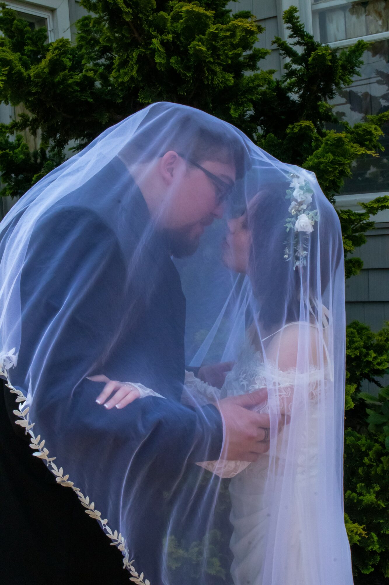 A couple under a wedding veil outdoors, with trees and a building in the background.