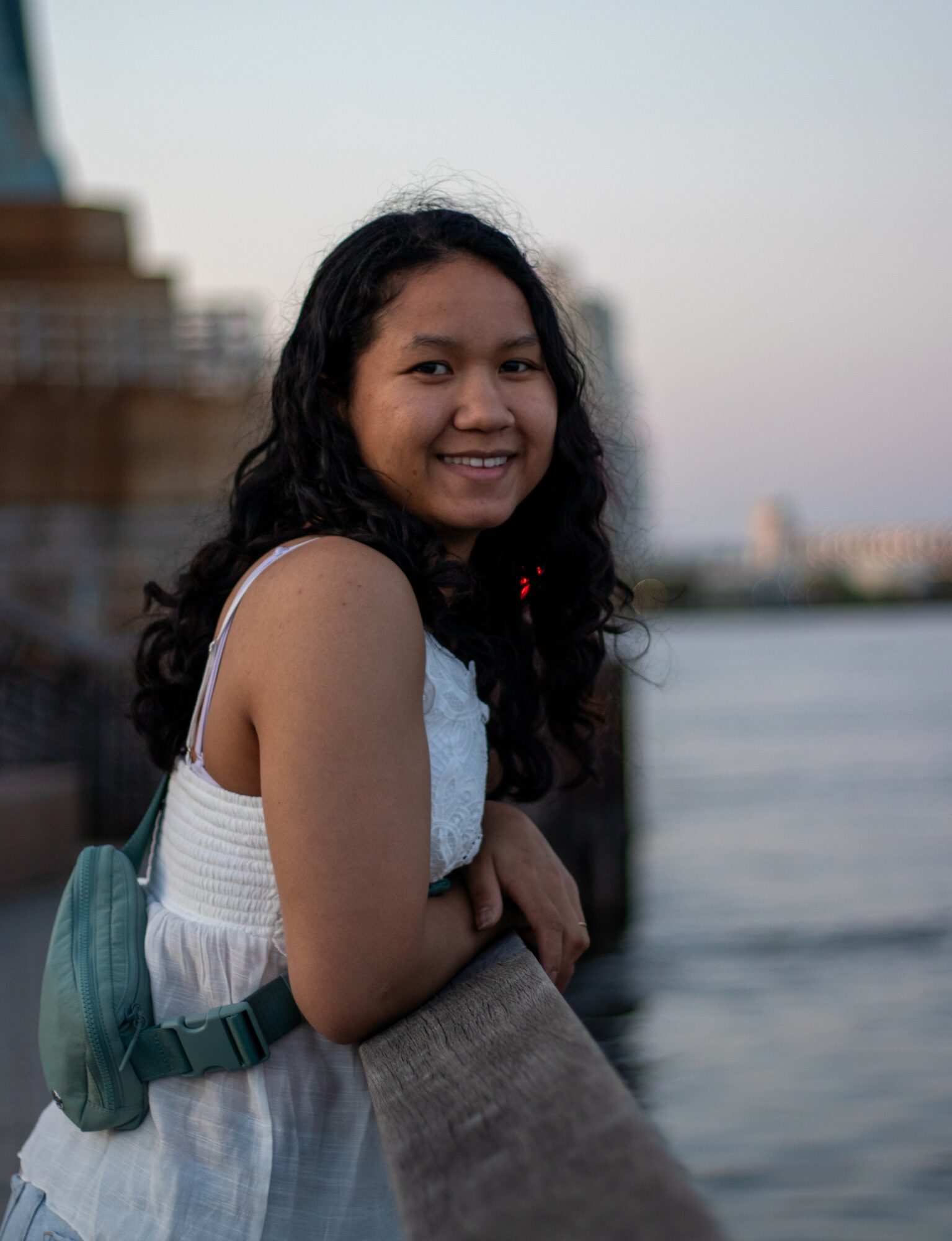 Smiling girl with long dark hair standing near water, wearing a white sleeveless top and a backpack.