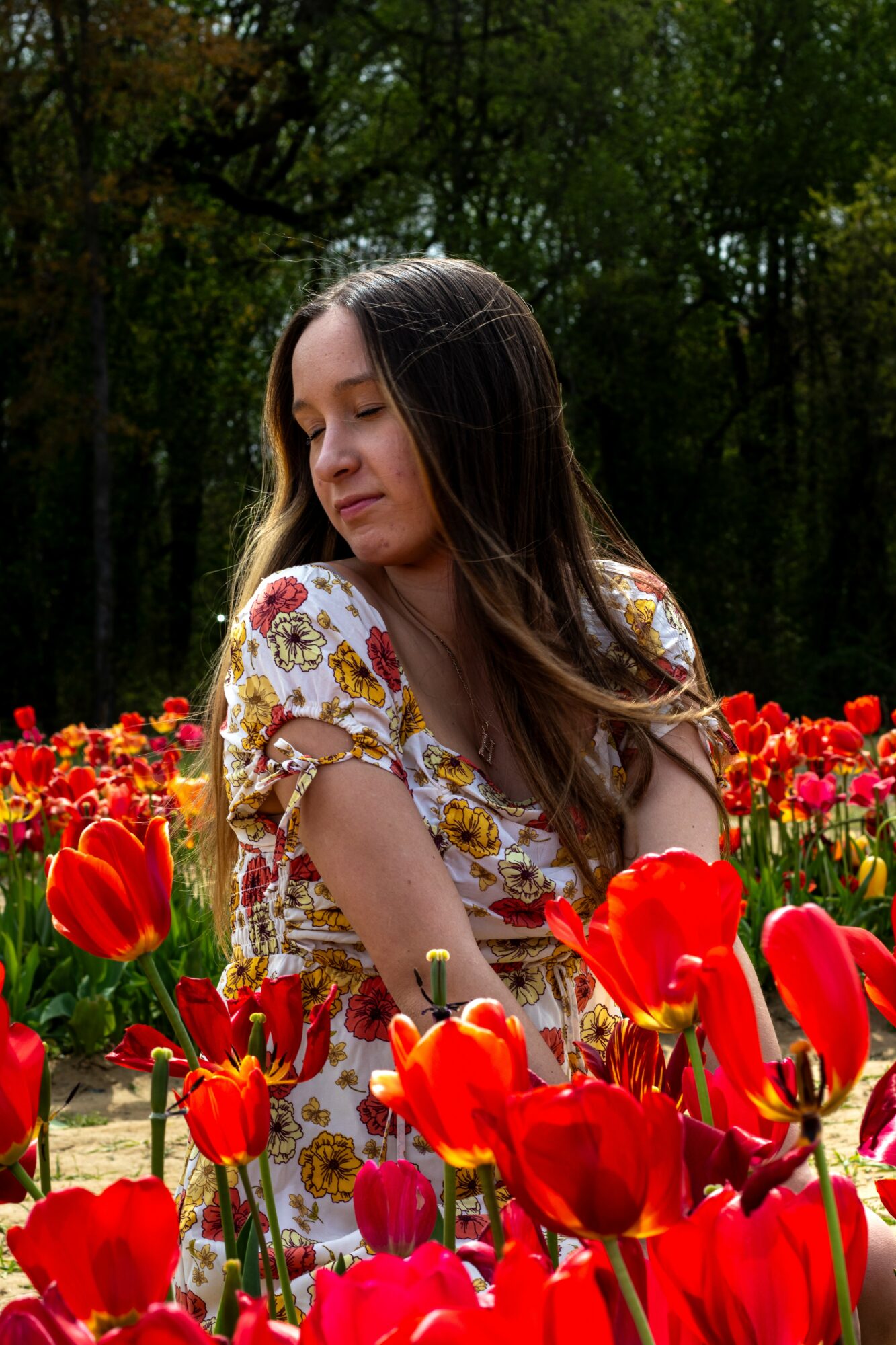 Young woman with long hair in a floral dress among red tulips outdoors, trees in background, eyes closed, serene expression.