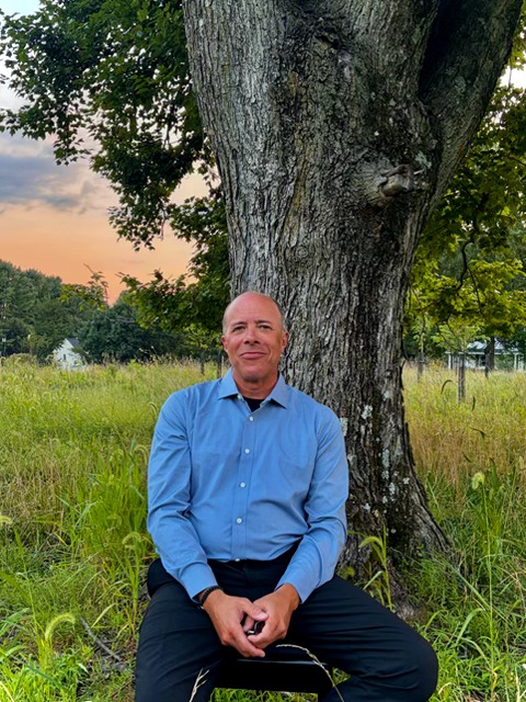Man sitting on grass in front of a large tree during sunset, wearing a blue shirt and black pants.