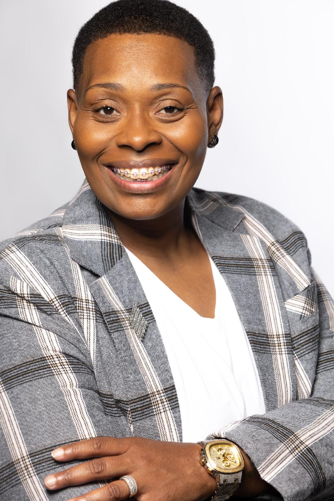 Portrait of a smiling woman with short hair, wearing a plaid blazer and a white top, crossing her arms, against a plain background.