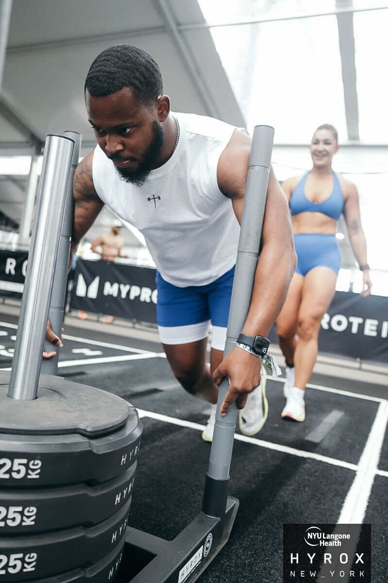 Man performing a push-up with weights at gym, woman in blue sportswear in background, bright indoor setting.