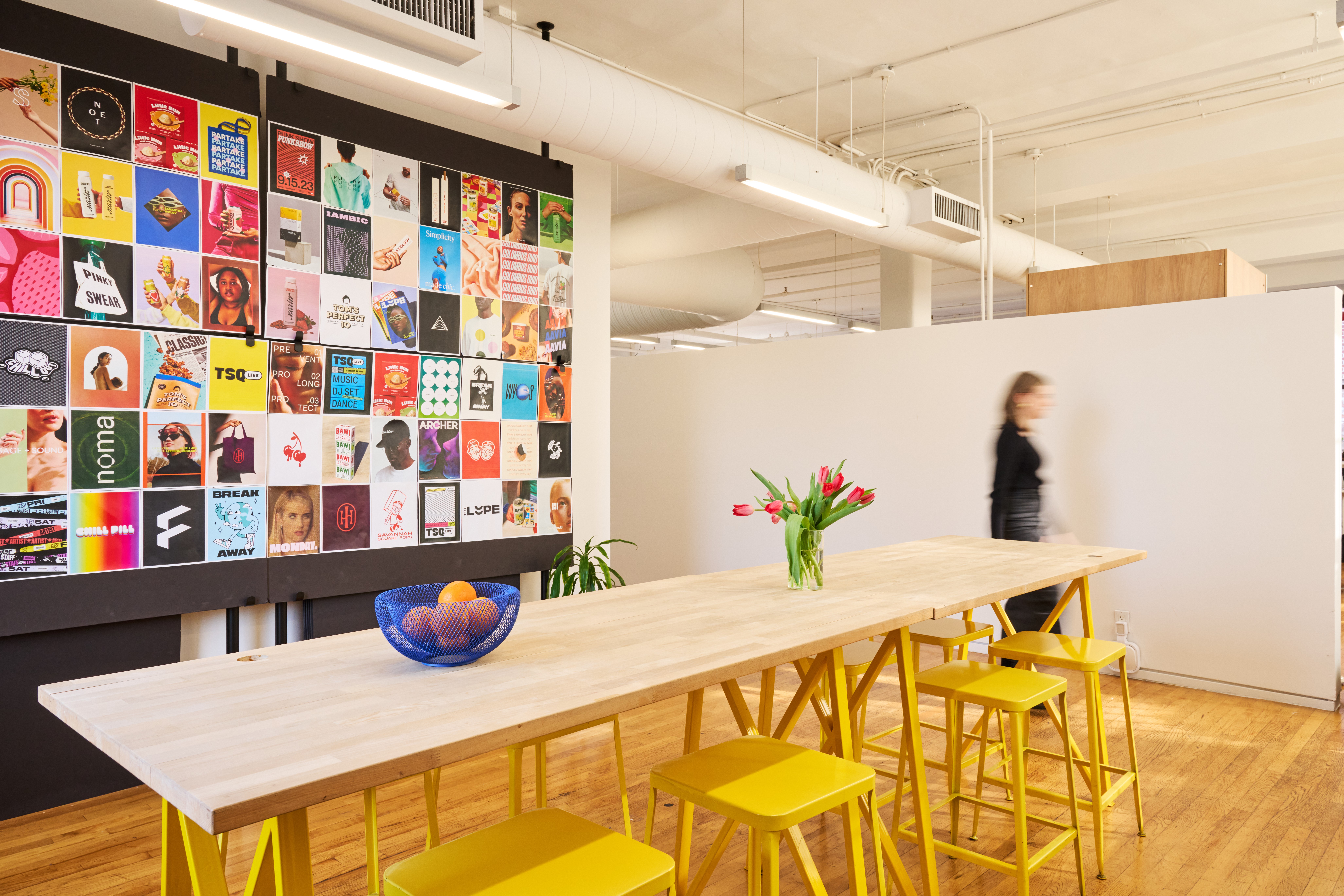 Long wooden table with yellow chairs, a bowl of fruit, and a vase with flowers in a modern room with artwork on the wall.
