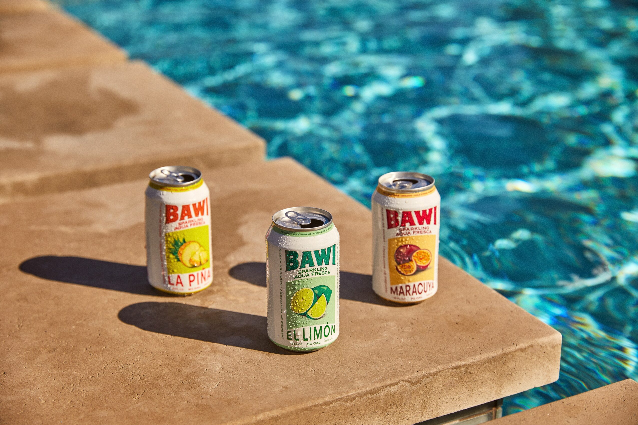Three cans of flavored drinks on poolside edge, with water in the background.