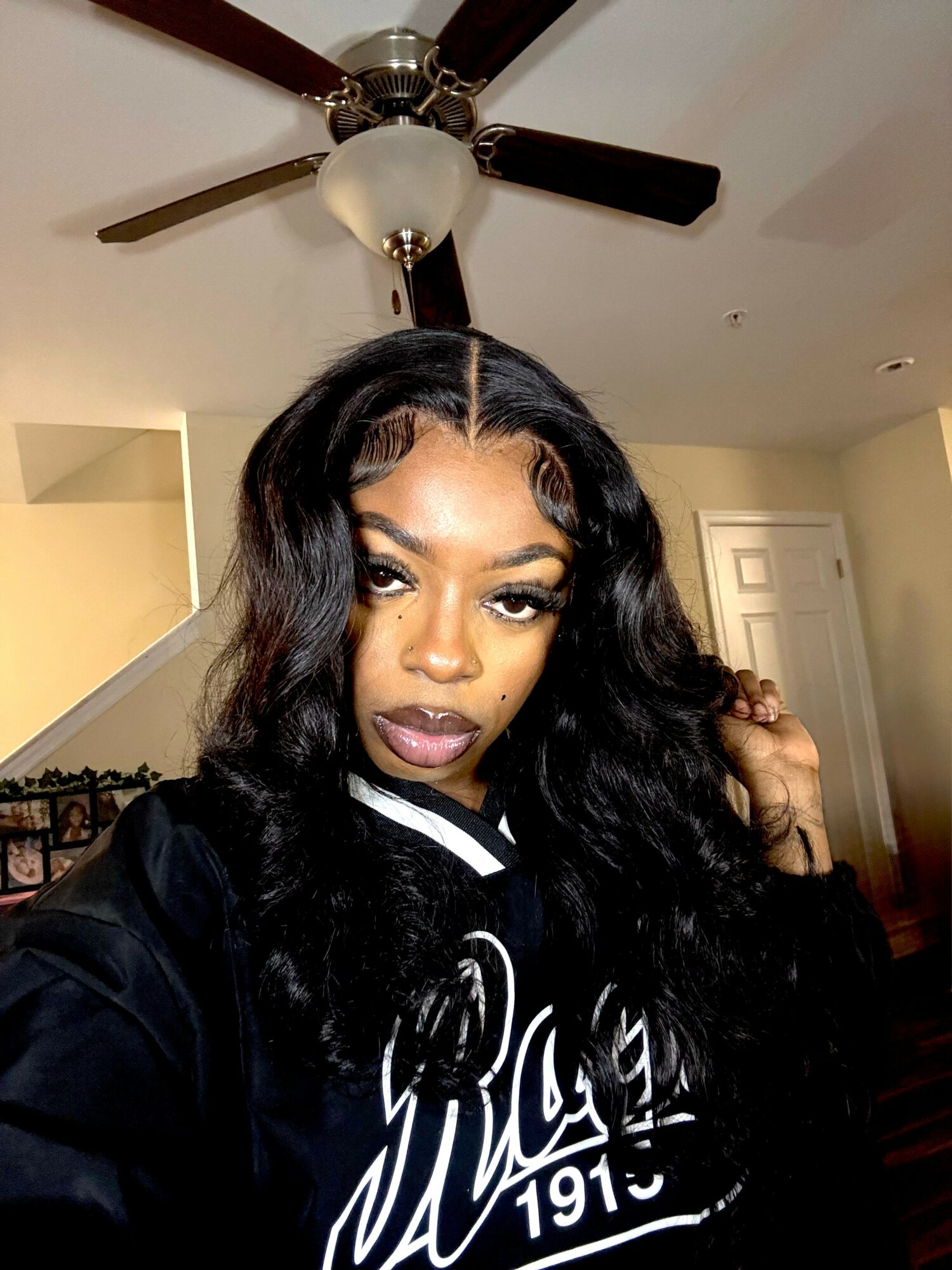 Young woman with long wavy hair taking a selfie indoors, ceiling fan above, neutral background.