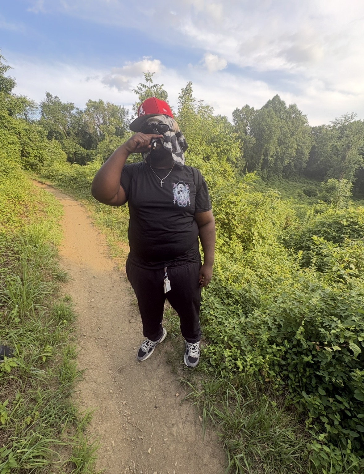 Person standing on a dirt path in a green outdoor area, taking a selfie with a phone, wearing a black shirt, black pants, and a red cap.