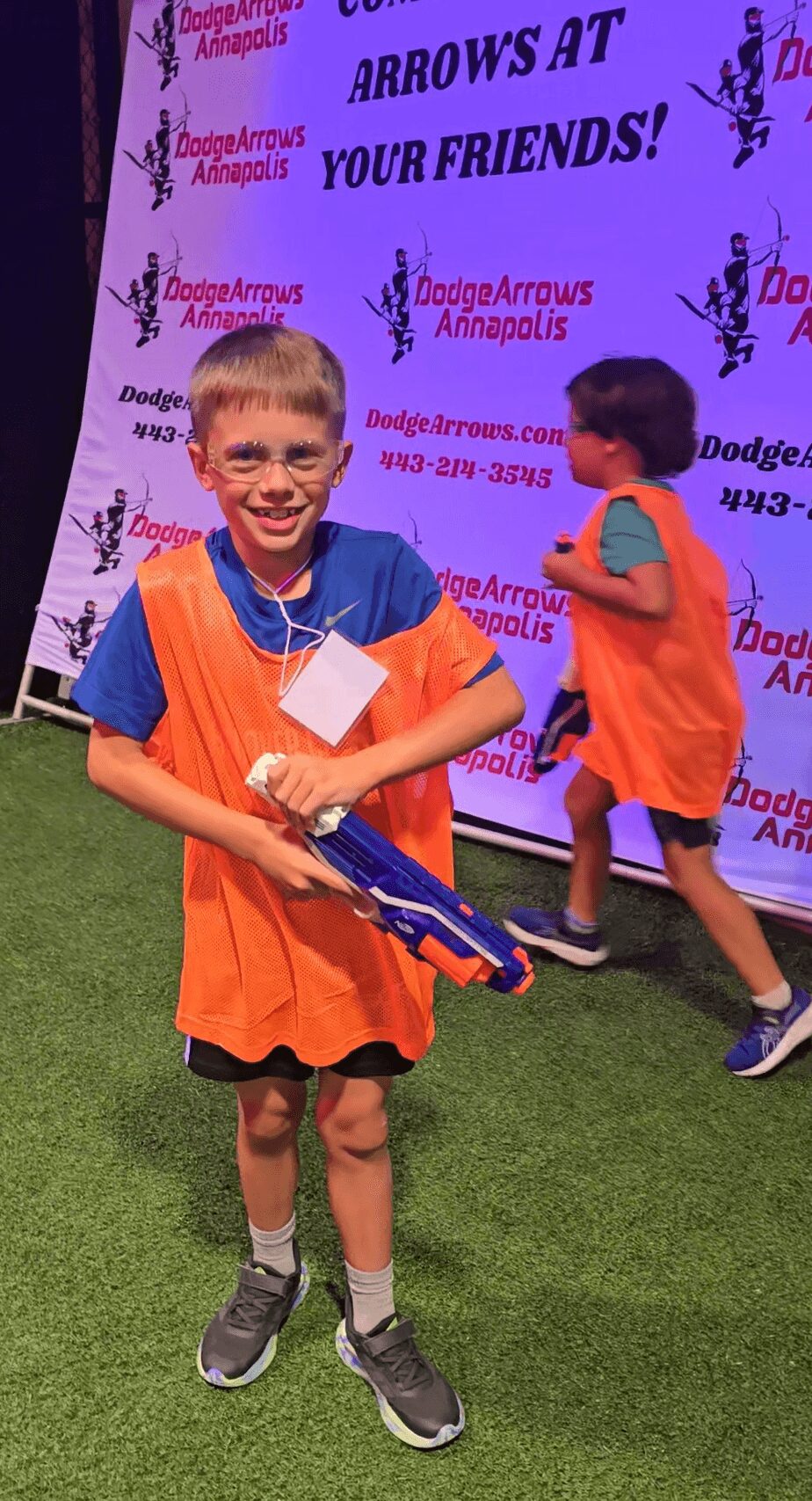 Child smiling with a medal and toy gun, standing on grass with a purple event backdrop behind.