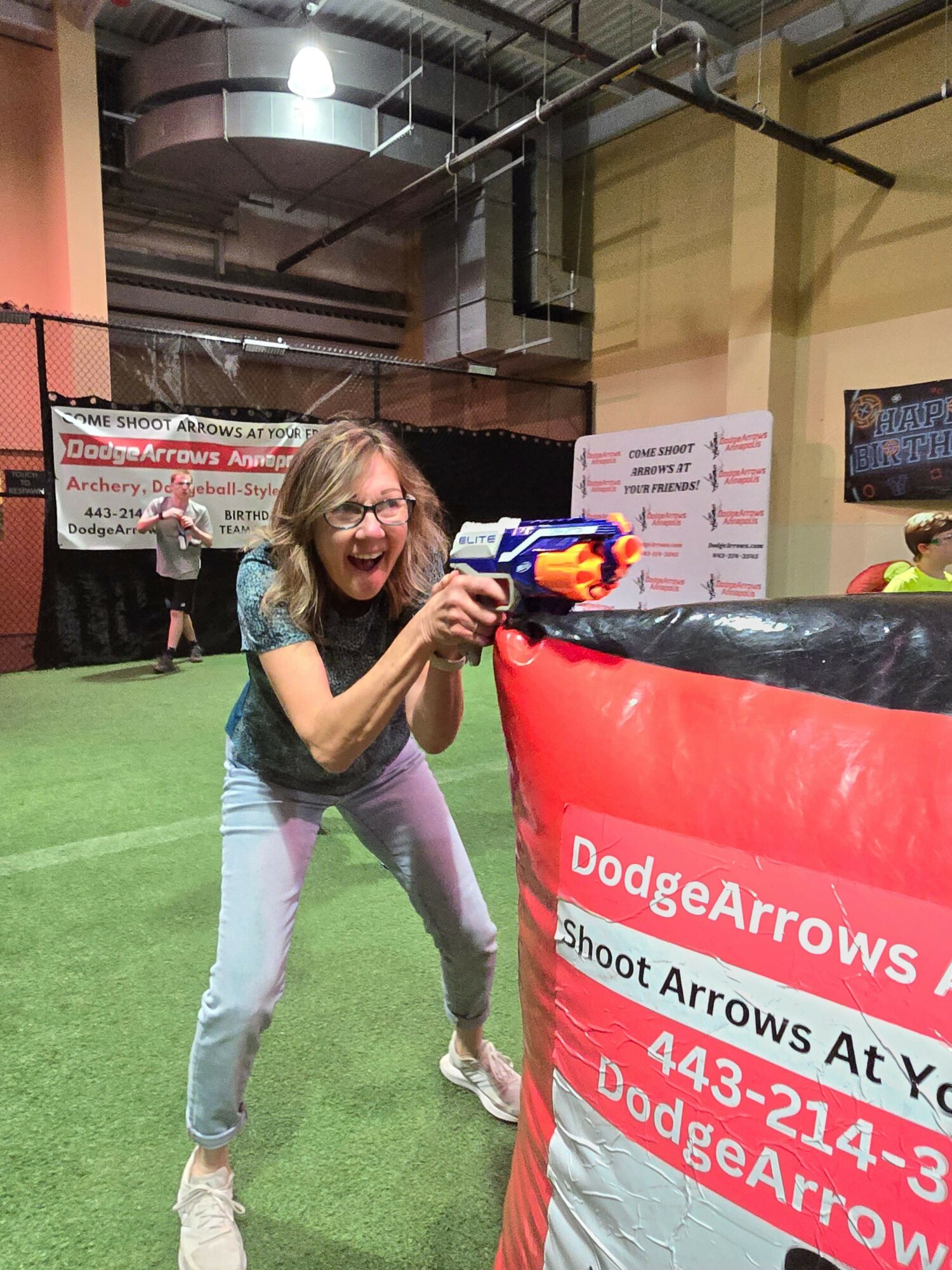 Woman holding a toy gun at an indoor event, standing on green artificial turf, with banners and signs in the background.