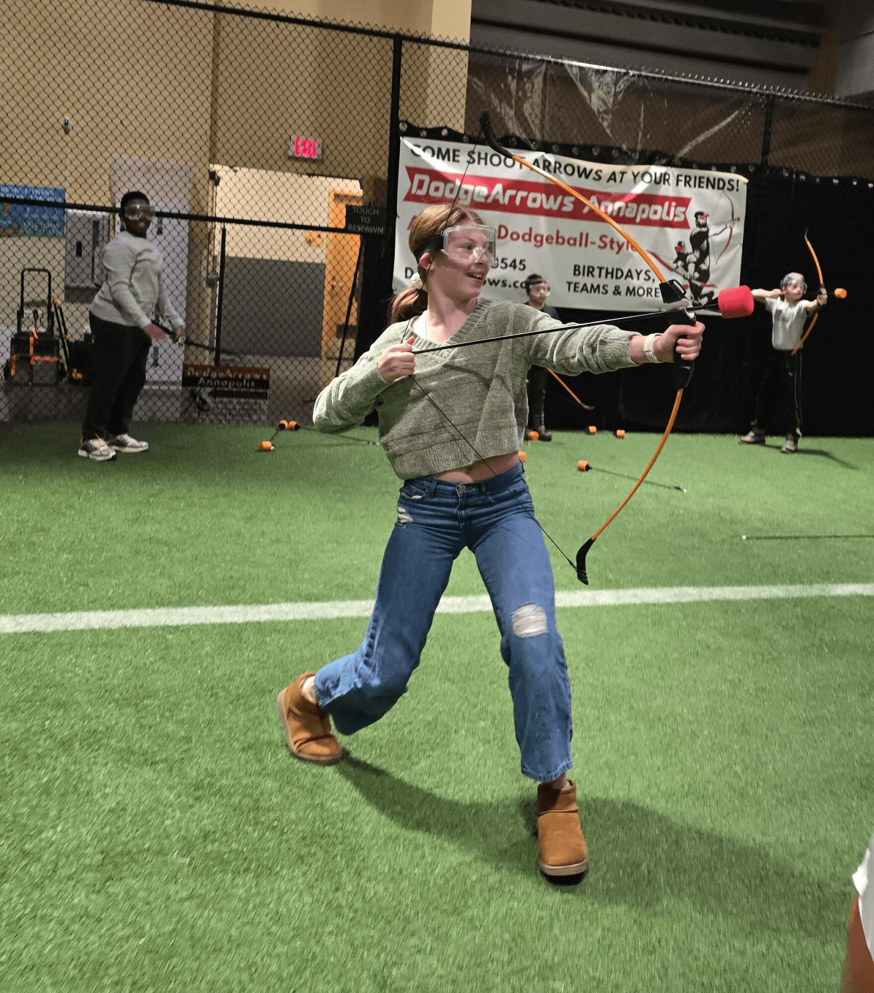 Person with glasses and pink hair practicing archery indoors on green turf, with others in the background near a netted area.