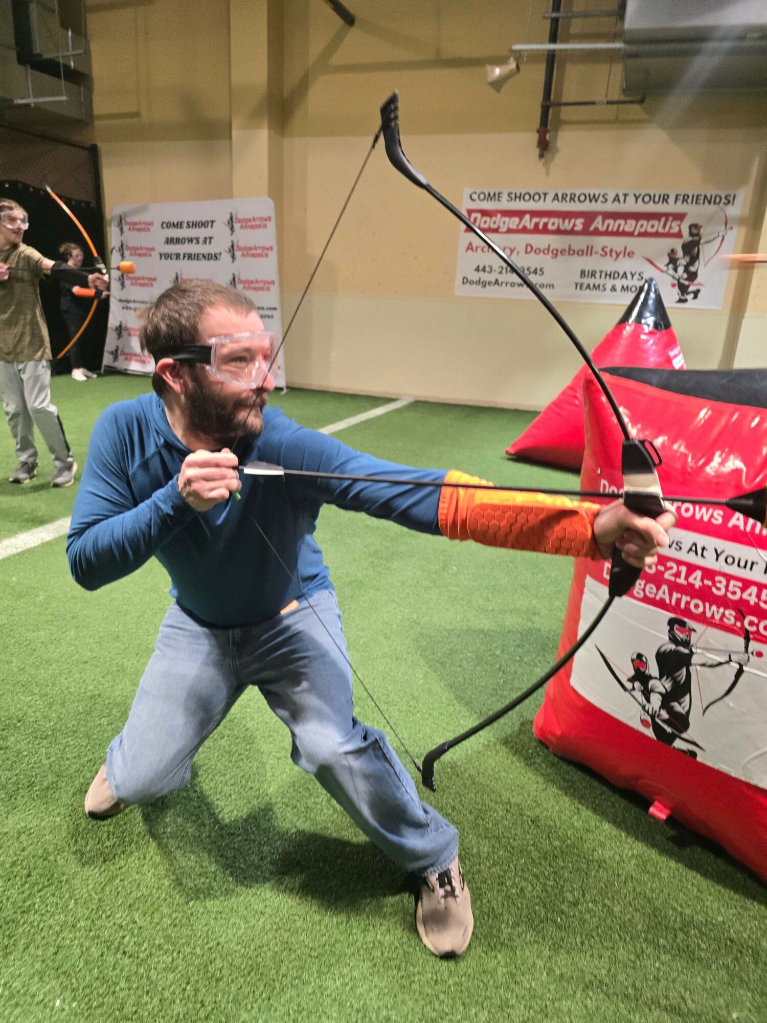 Person practicing archery indoors, drawing a bow with an arrow, on green artificial turf, with banners in background.