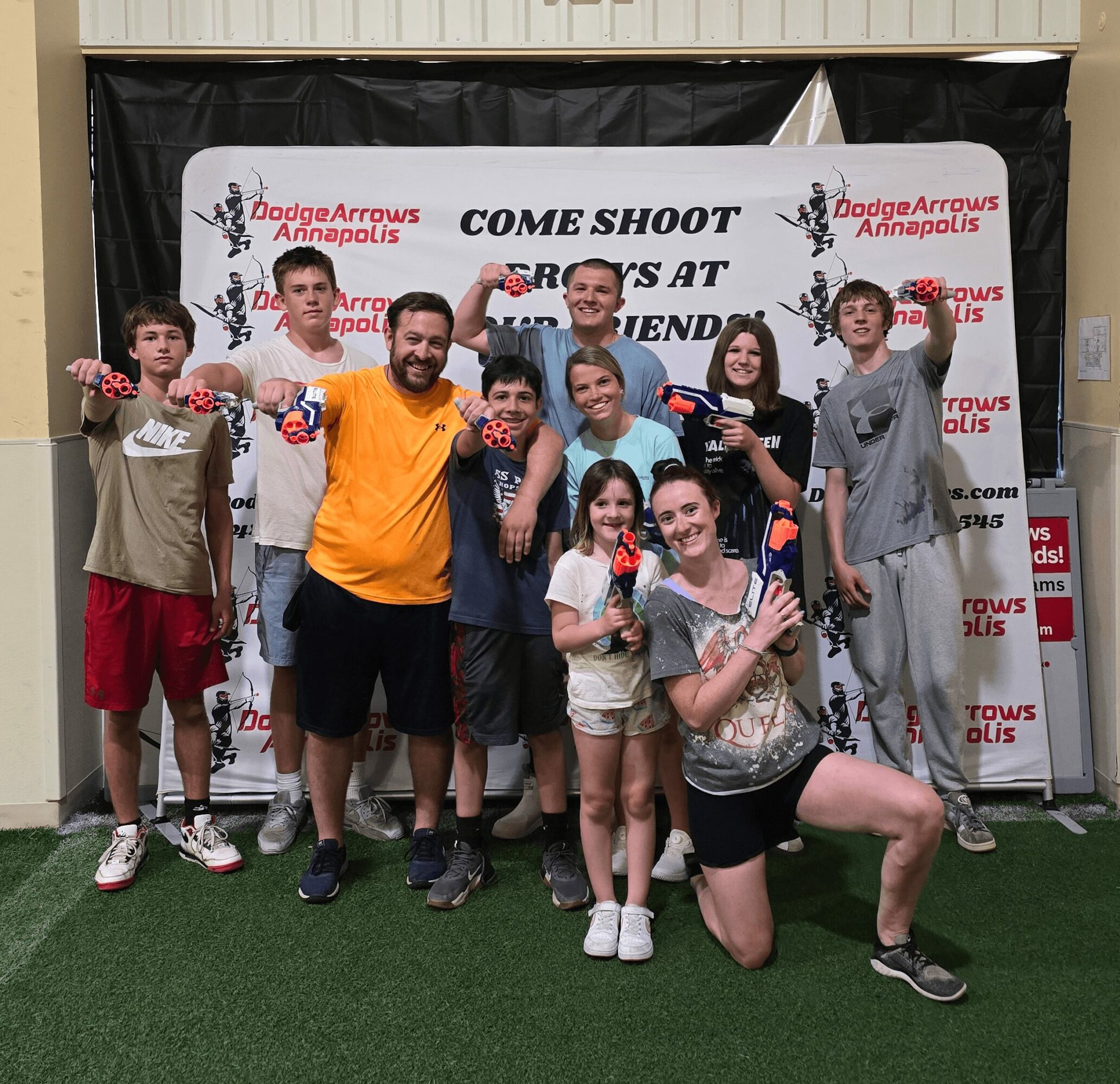 Group of children and adults holding foam dart guns, smiling, in front of a banner at an indoor event.