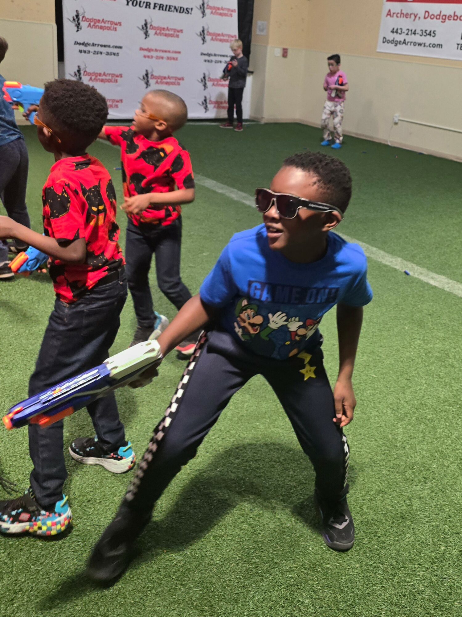 Children playing indoors on green artificial turf, one boy in sunglasses and a blue shirt crouching, others in red shirts nearby.