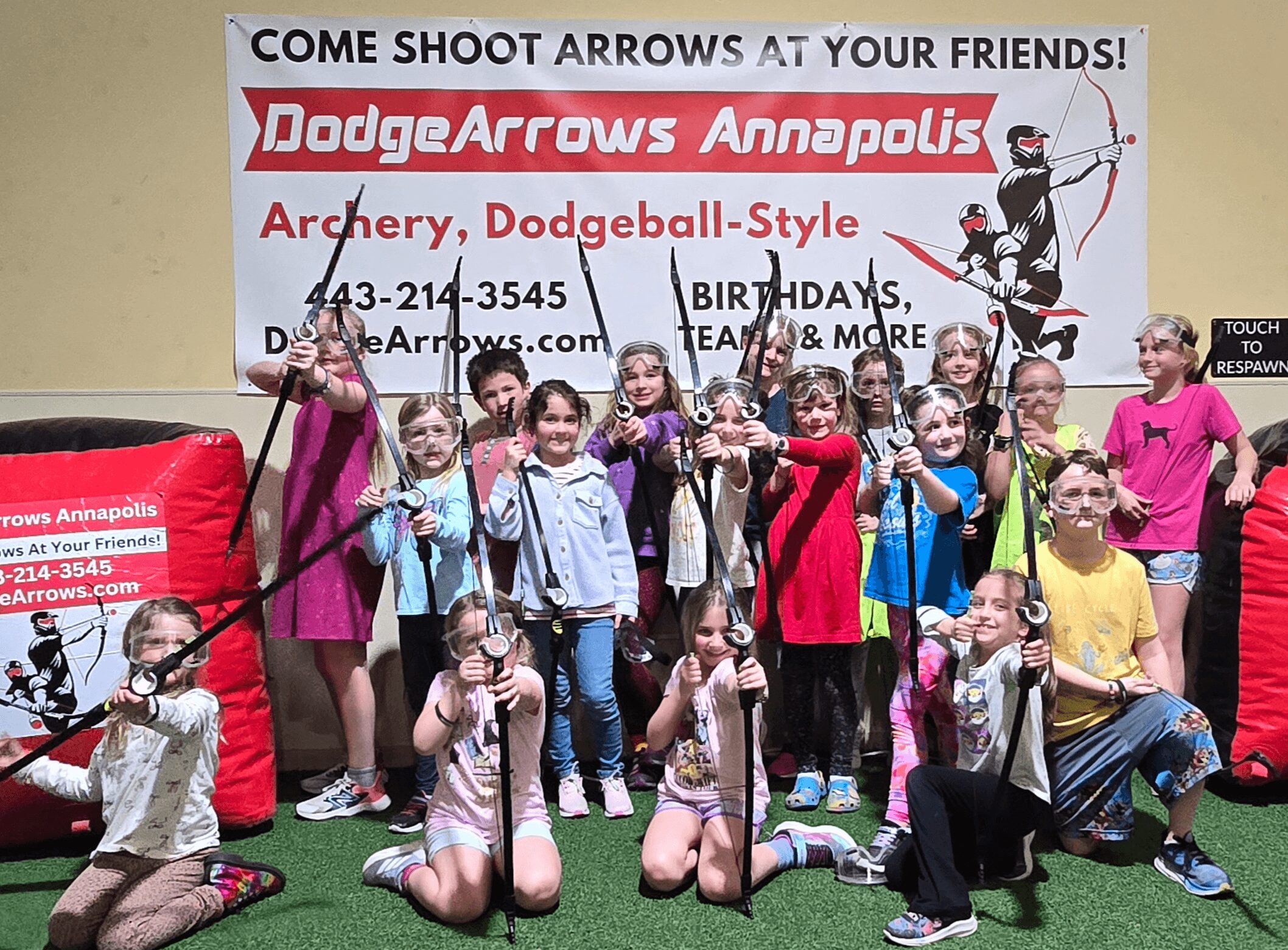 Group of children and adults holding bows and arrows, posing in front of a sign for archery activities.
