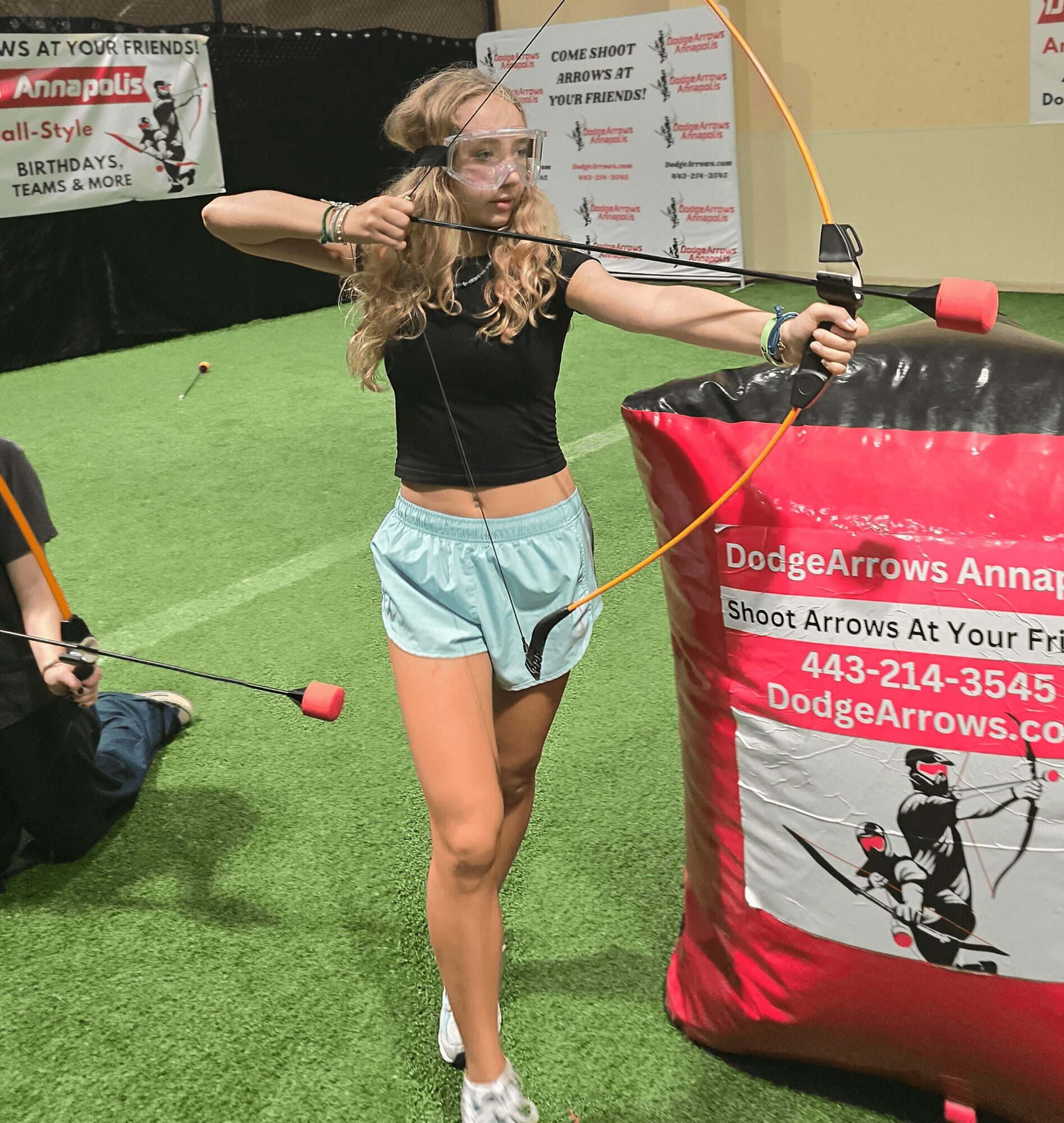 Young girl with curly hair aiming a bow and arrow at a target indoors, wearing a black top and light blue shorts.