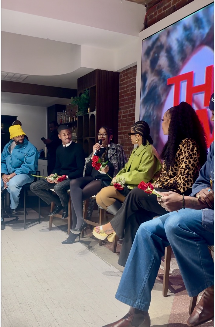 Five people sit on chairs in a room, holding flowers, with a large screen displaying a logo behind them.