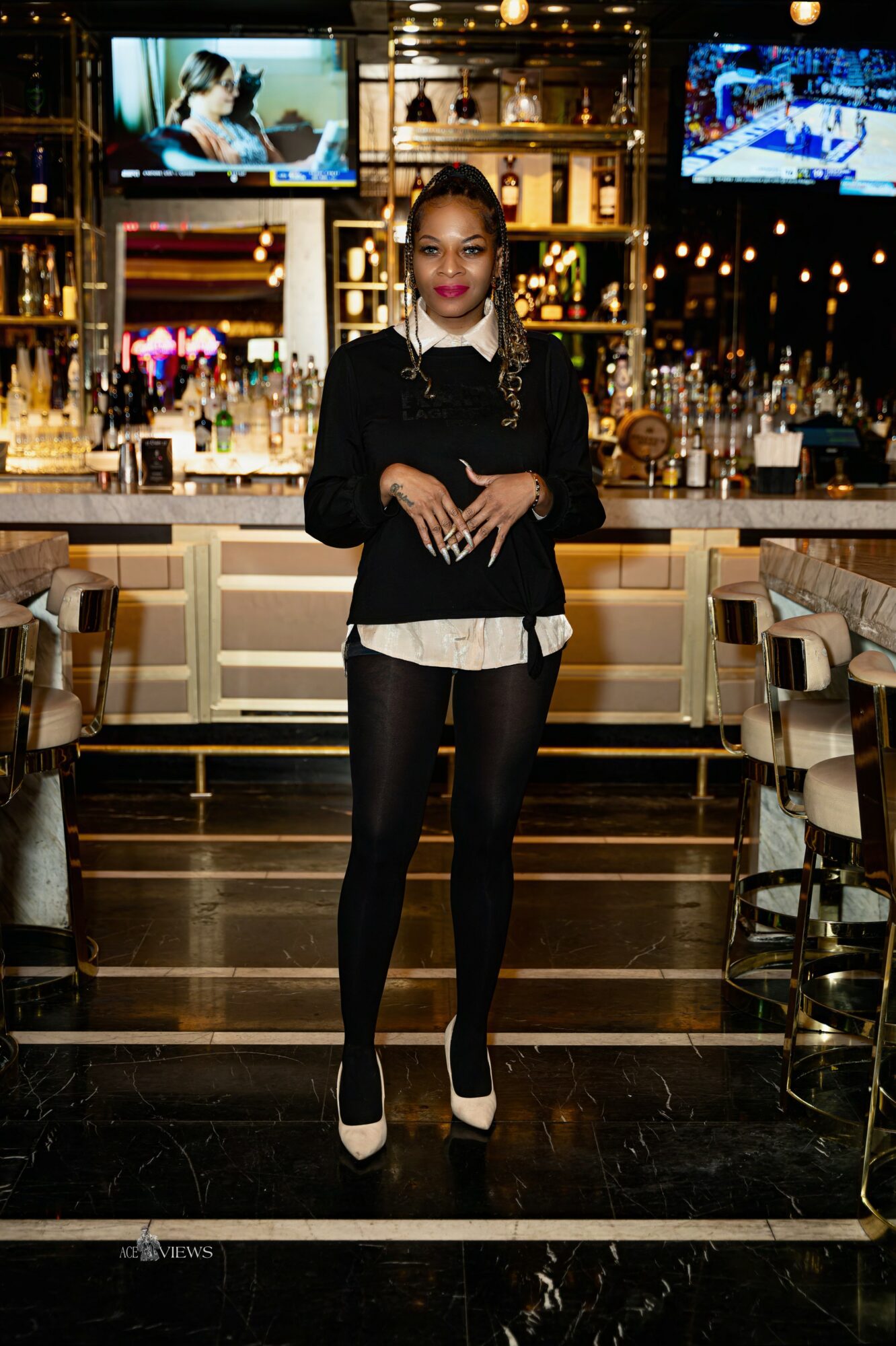 Woman standing in a bar or restaurant with TVs and bar shelves behind her, wearing a black top, white shirt, and black pants.