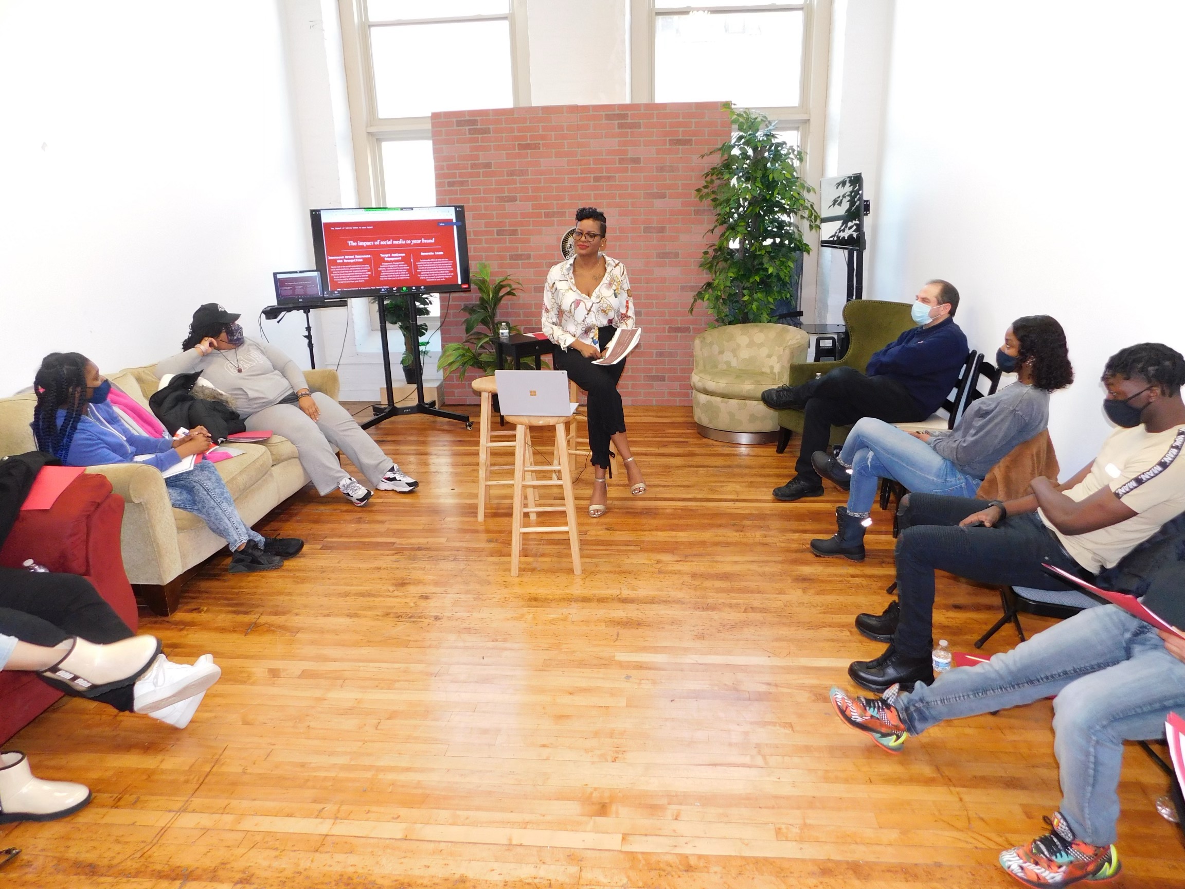 Person standing in front of a group in a room with wooden floor, brick wall, and large windows, giving a presentation.