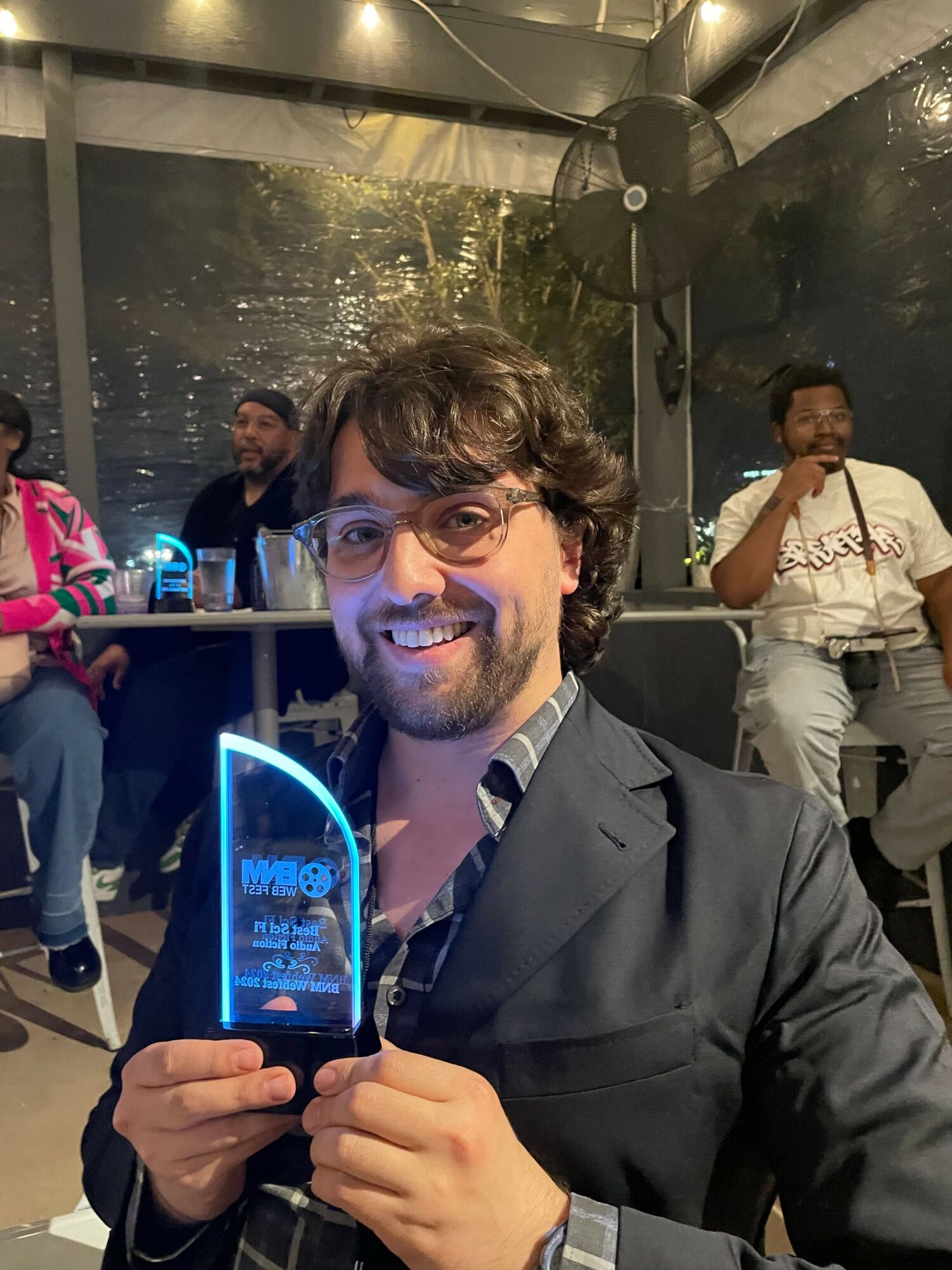 Man with dark hair and beard smiling, holding a phone with a blue glow, in a lively indoor setting.