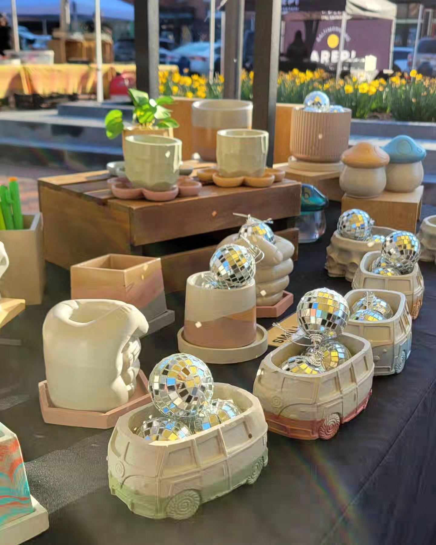 Display of ceramic and decorative items on a table outdoors, with flowers and a building in the background.