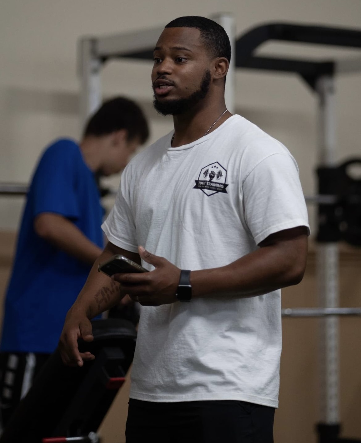 Man standing indoors holding a phone, wearing a white T-shirt with a logo, with a person in blue in the background.