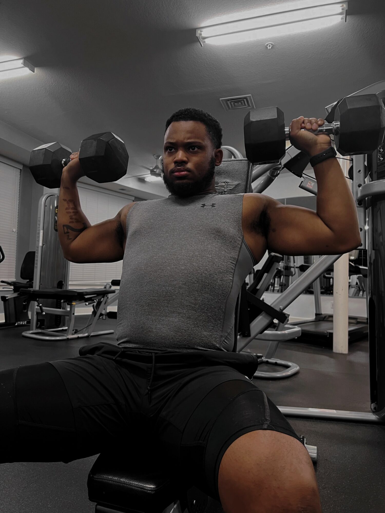 Man lifting dumbbells in gym, seated with arms raised, wearing sleeveless shirt, focused expression, gym equipment in background.