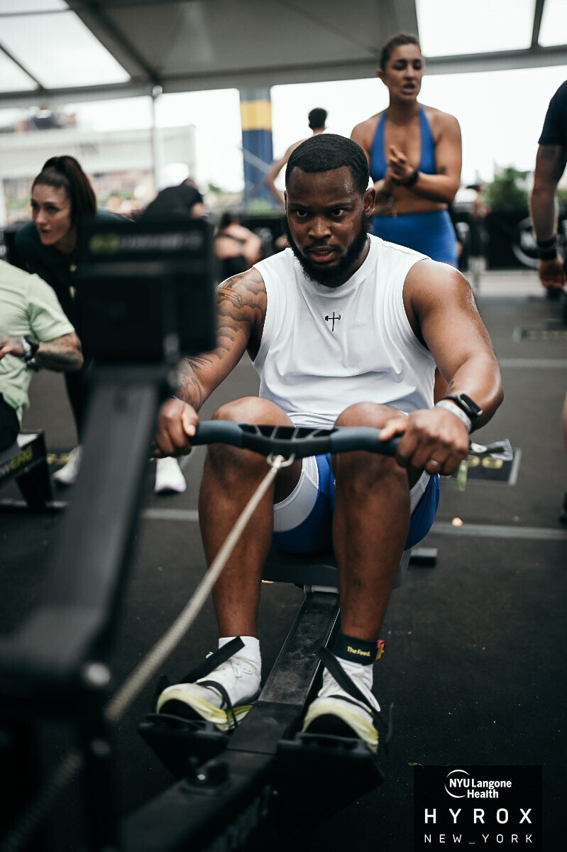 Man using rowing machine at gym with two women in background, one standing and one sitting, in a fitness setting.