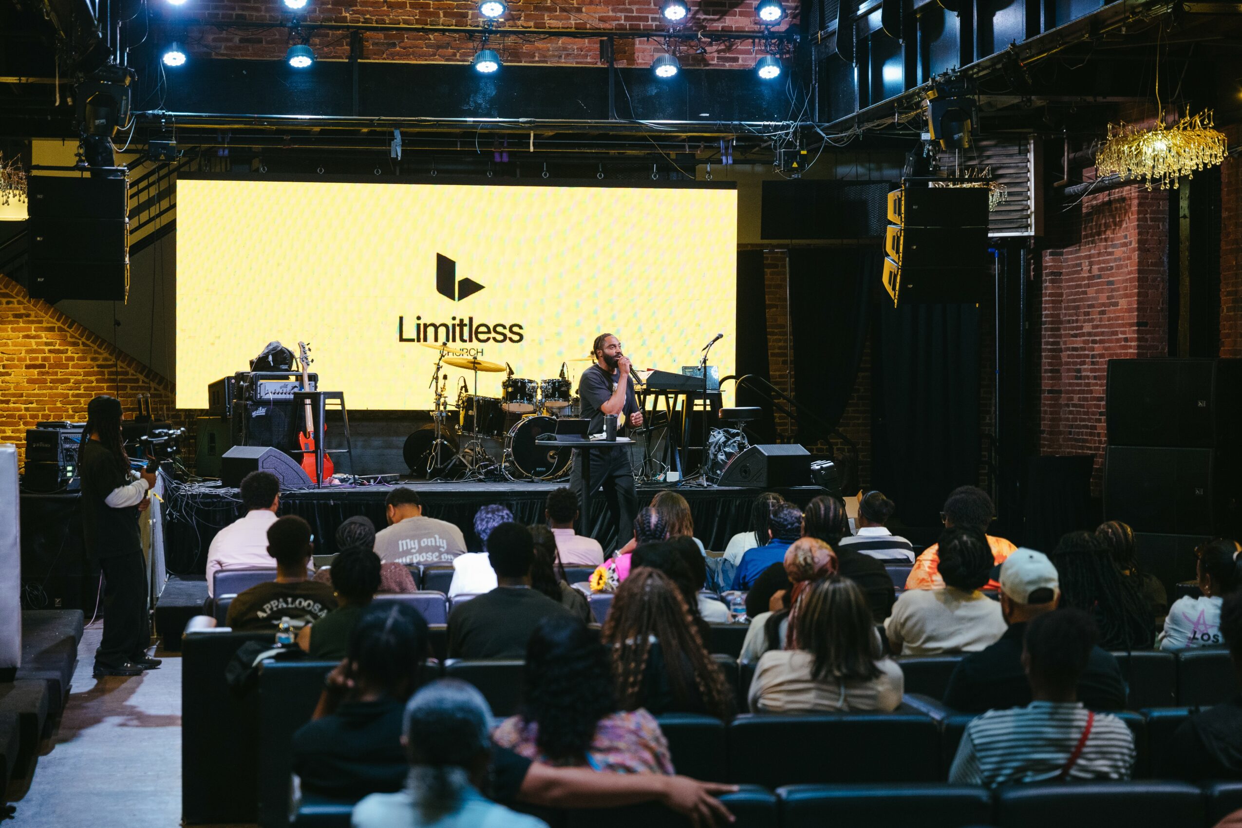 Audience watching a live performance on stage with a large yellow screen displaying 'Limitless' logo.