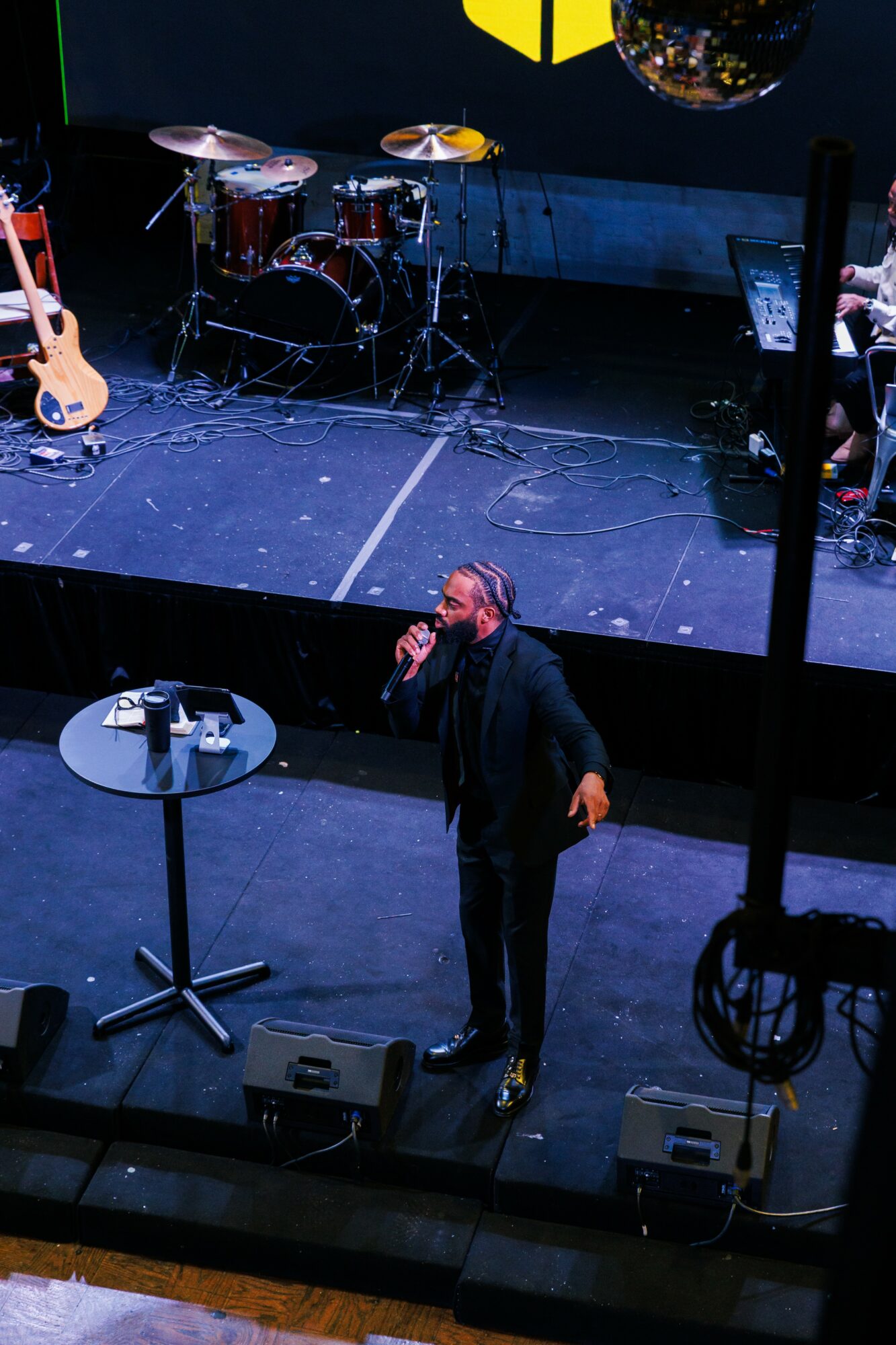 Performer on stage with musical instruments, microphone, and a person speaking into a microphone, stage setup visible.