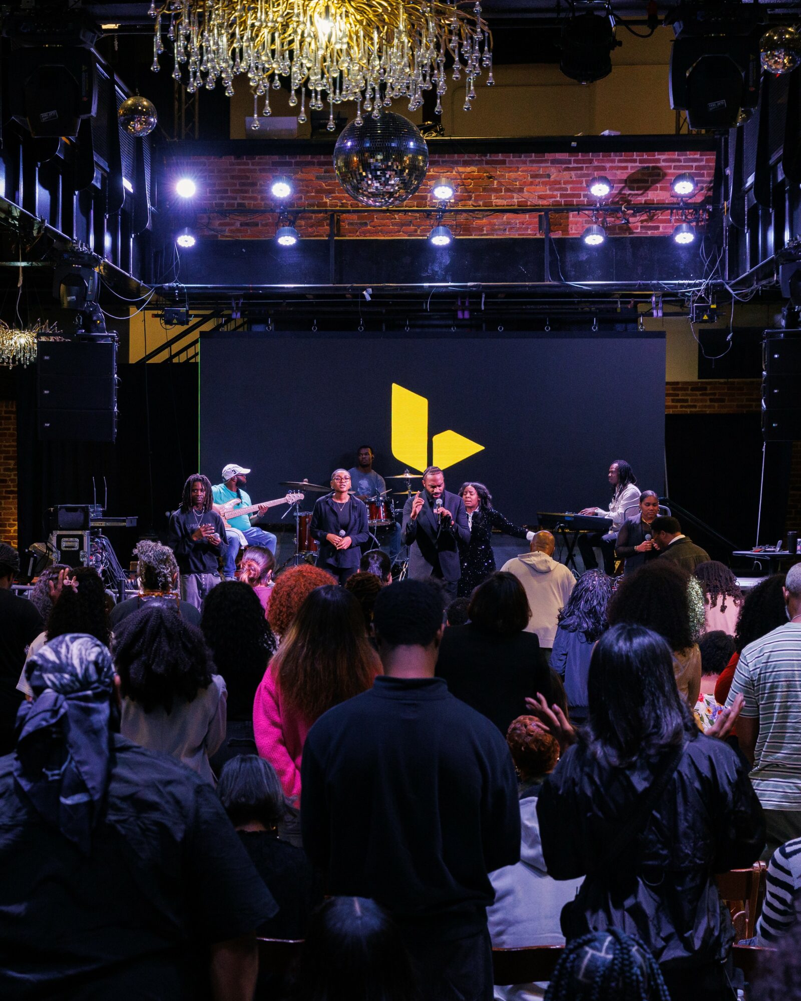 People gathered in a venue watching a panel or performance on stage with a large screen and lighting, chandelier overhead.