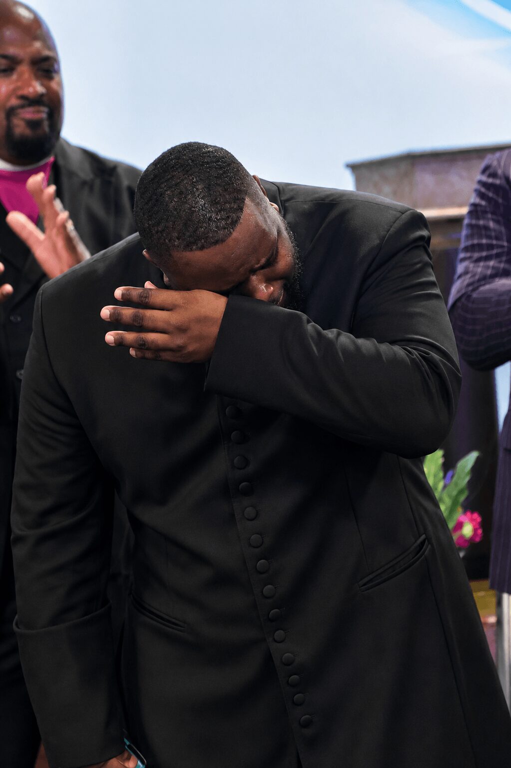 Person in black suit covering face with arm, emotional moment, others in background, outdoor setting