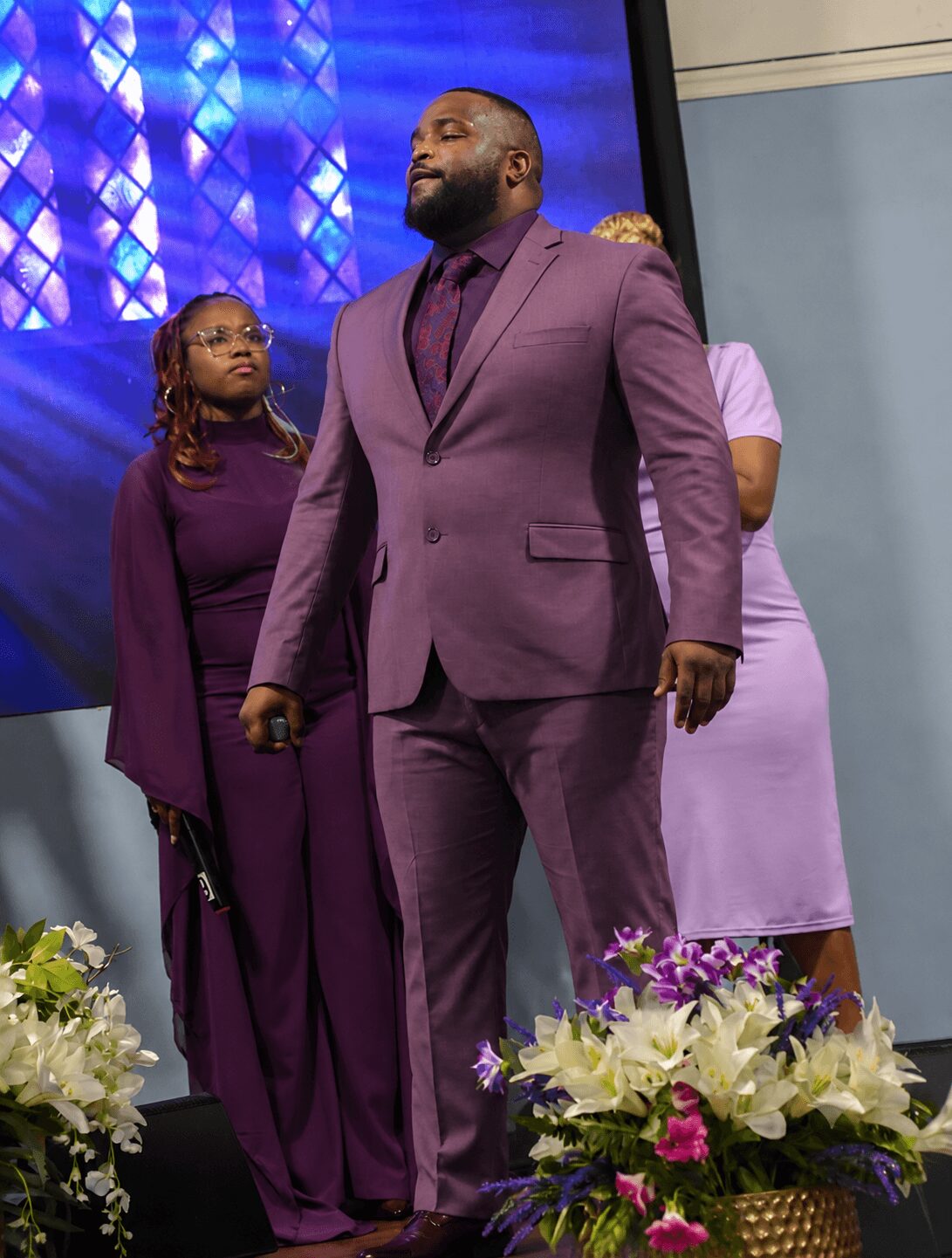Three people on stage, one man in a purple suit in front, two women behind him, flowers in foreground.