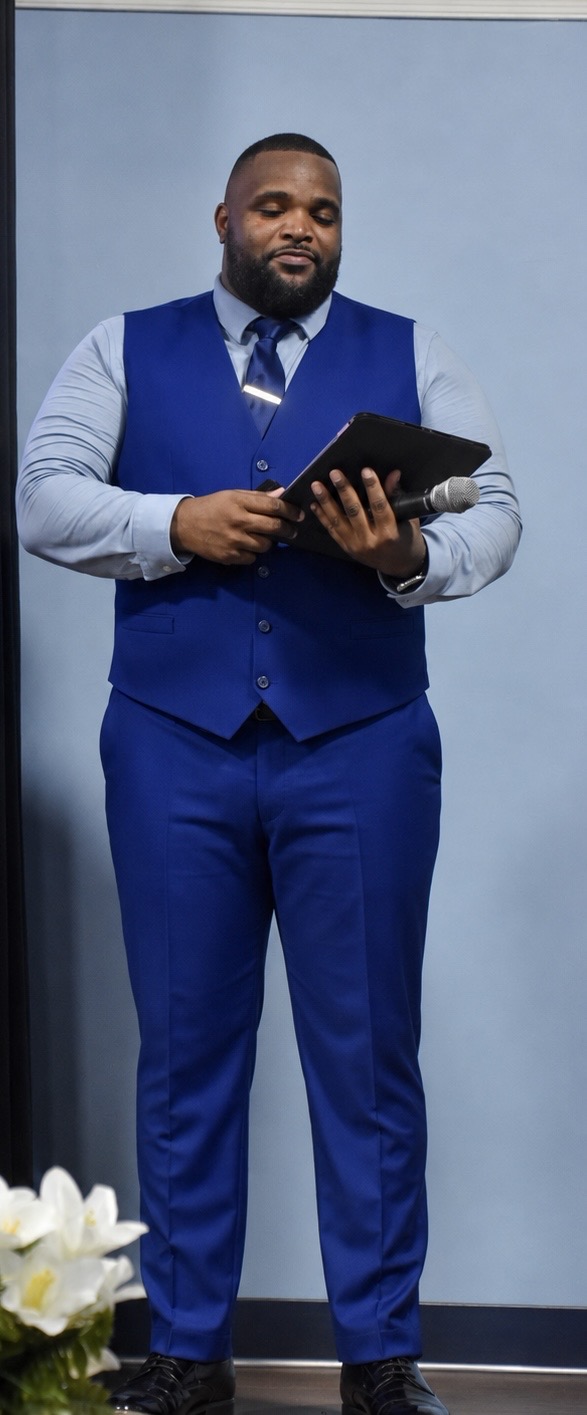 Man in blue suit holding a tablet, standing against a light blue wall.