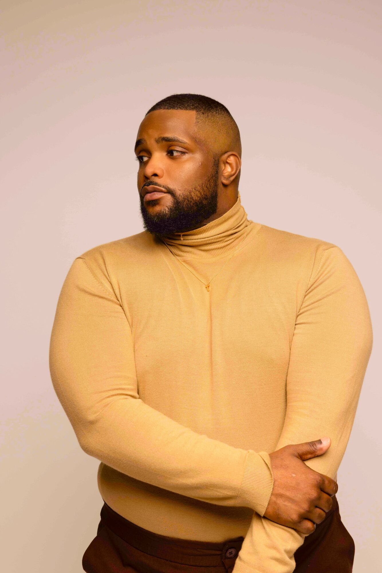Man with a beard and short hair wearing a beige turtleneck, looking to the side, arms crossed.