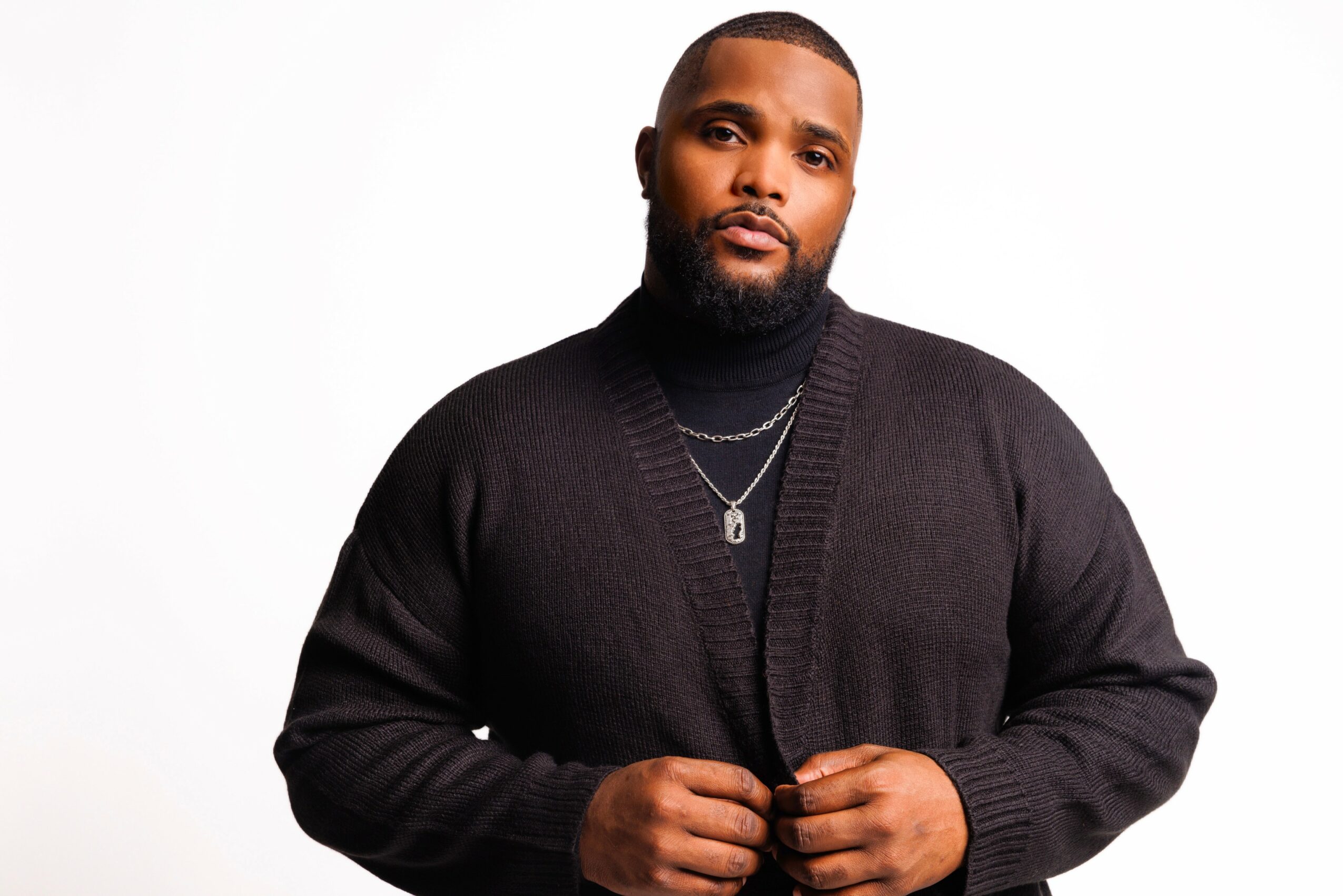 Man with a beard wearing a black cardigan and silver necklace, standing against a white background.