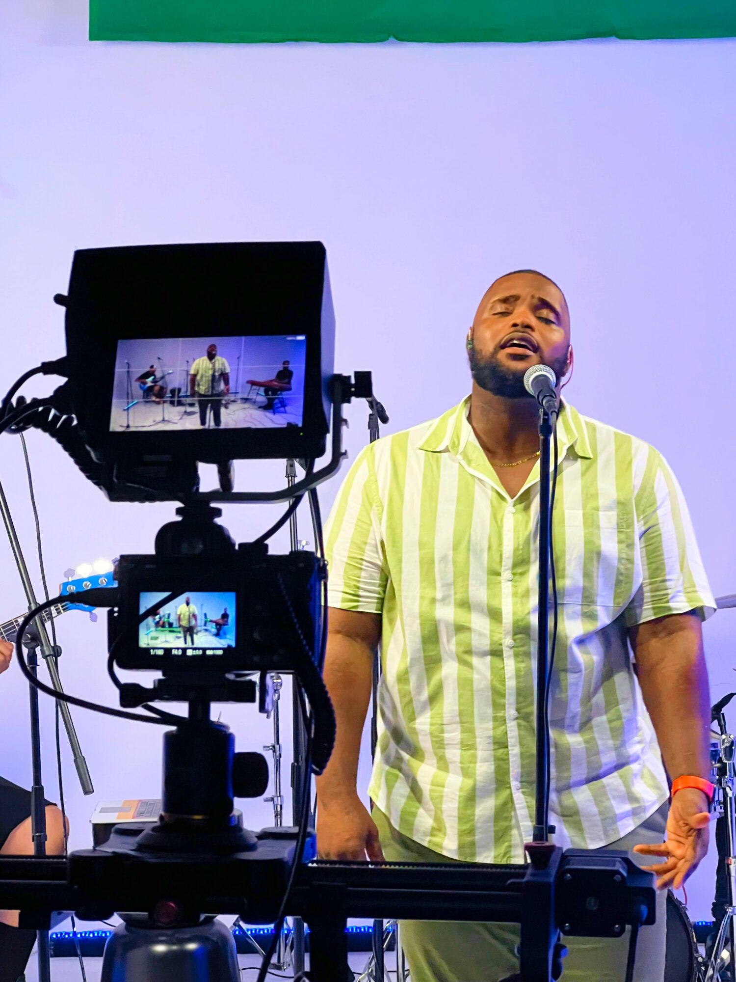 Man singing into microphone on stage with camera recording him, wearing a yellow and white striped shirt.
