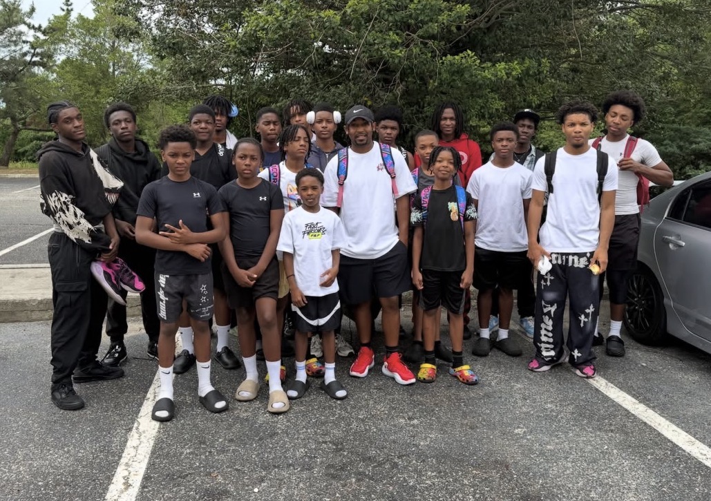 Group of young people and children standing outdoors on a parking lot with trees in background.