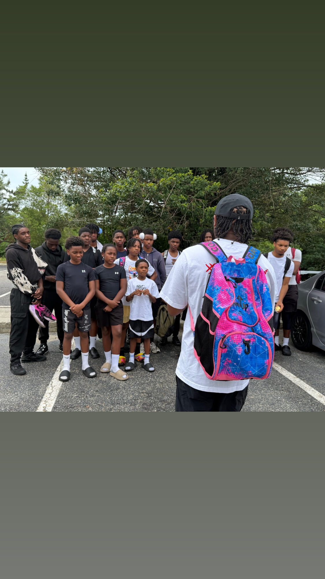 Group of people standing outdoors in a parking lot, with trees in the background, facing a person with a colorful backpack.