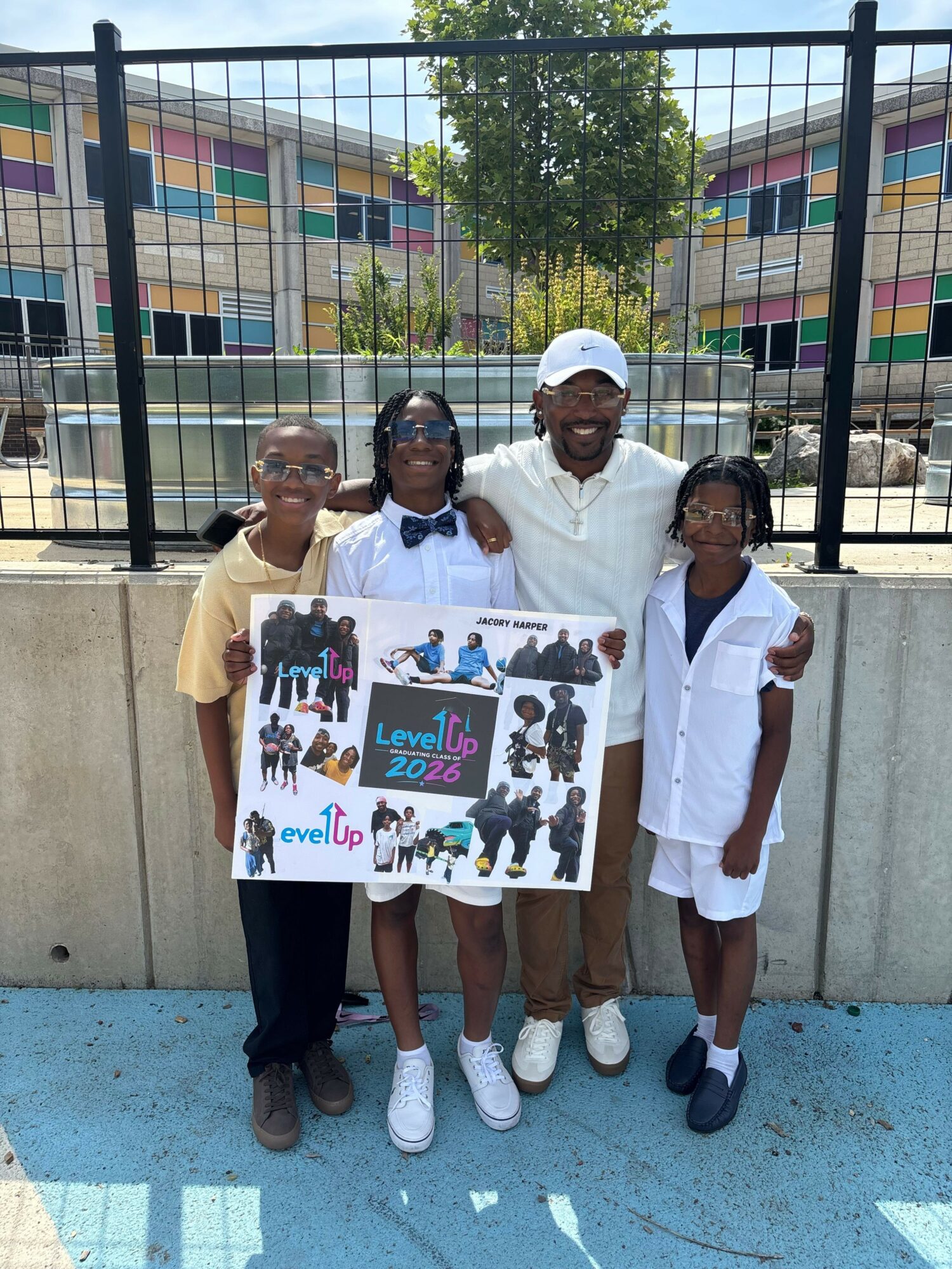 Four people holding a colorful poster with text and images, standing outdoors near a fence and colorful building.