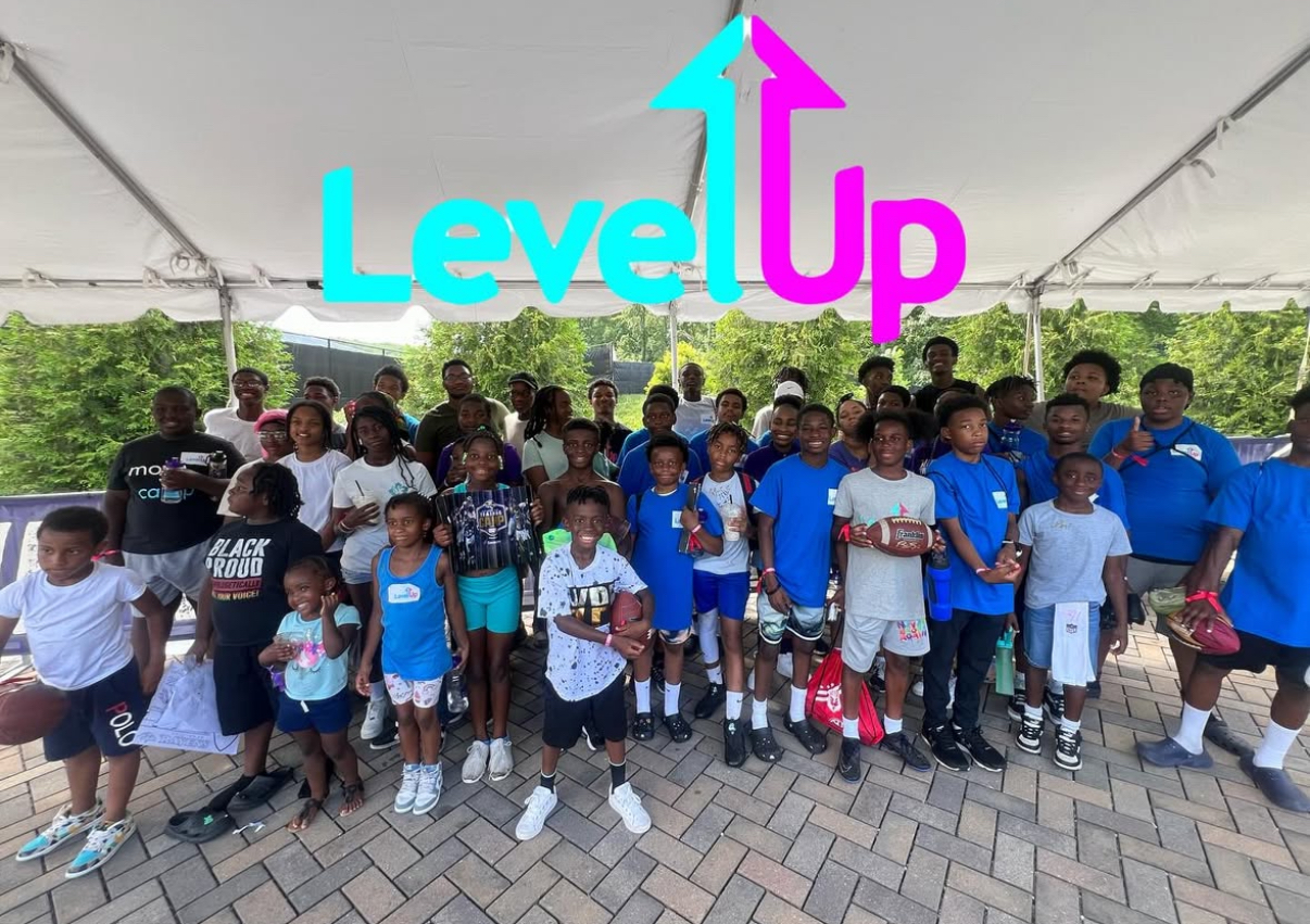 Group of children and adults outdoors under a tent, with a colorful 'Level Up' sign above them.