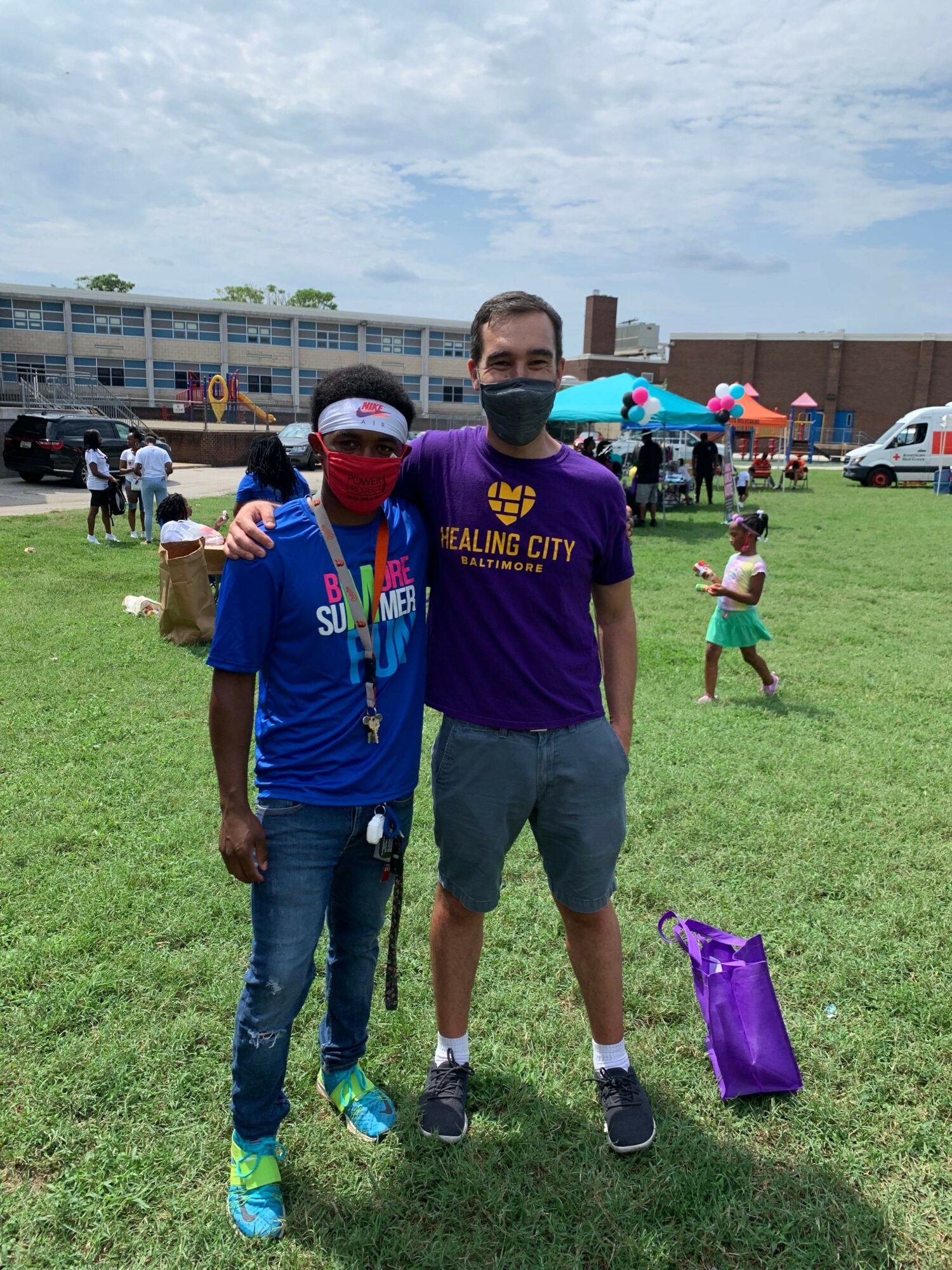 Two people standing on grass, one wearing a purple shirt and the other in a blue shirt, with children and tents in background.