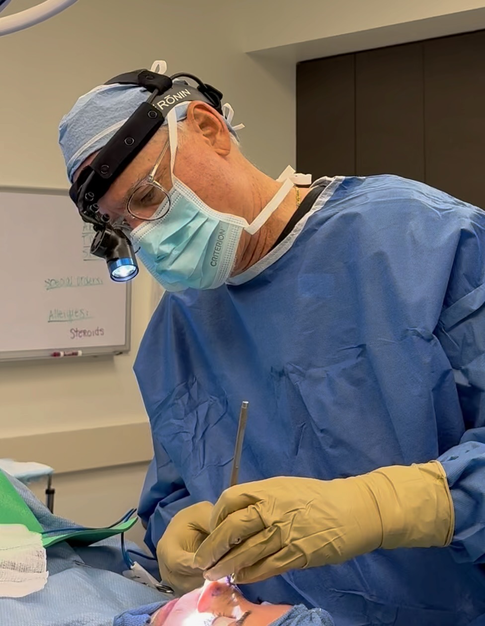 Surgeon in blue scrubs, mask, and magnifying glasses performs a procedure in an operating room.