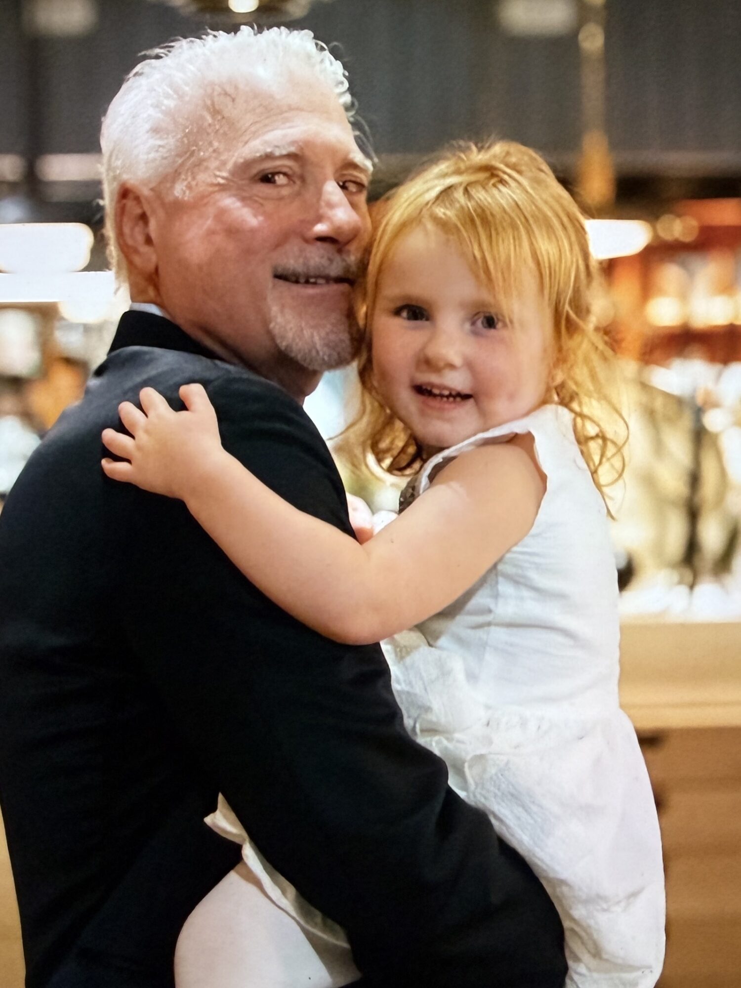 An elderly man with white hair and a beard holding a young girl with red hair, both smiling warmly indoors.