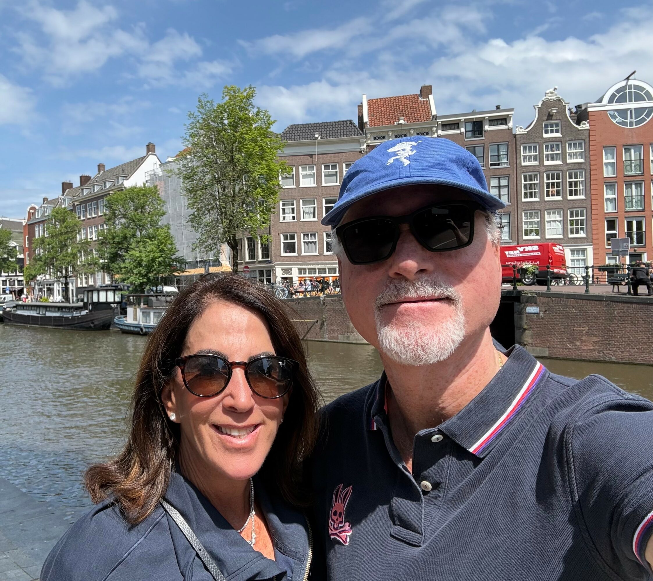 A smiling man and woman wearing sunglasses and hats taking a selfie by a canal with historic buildings and a blue sky in the background.