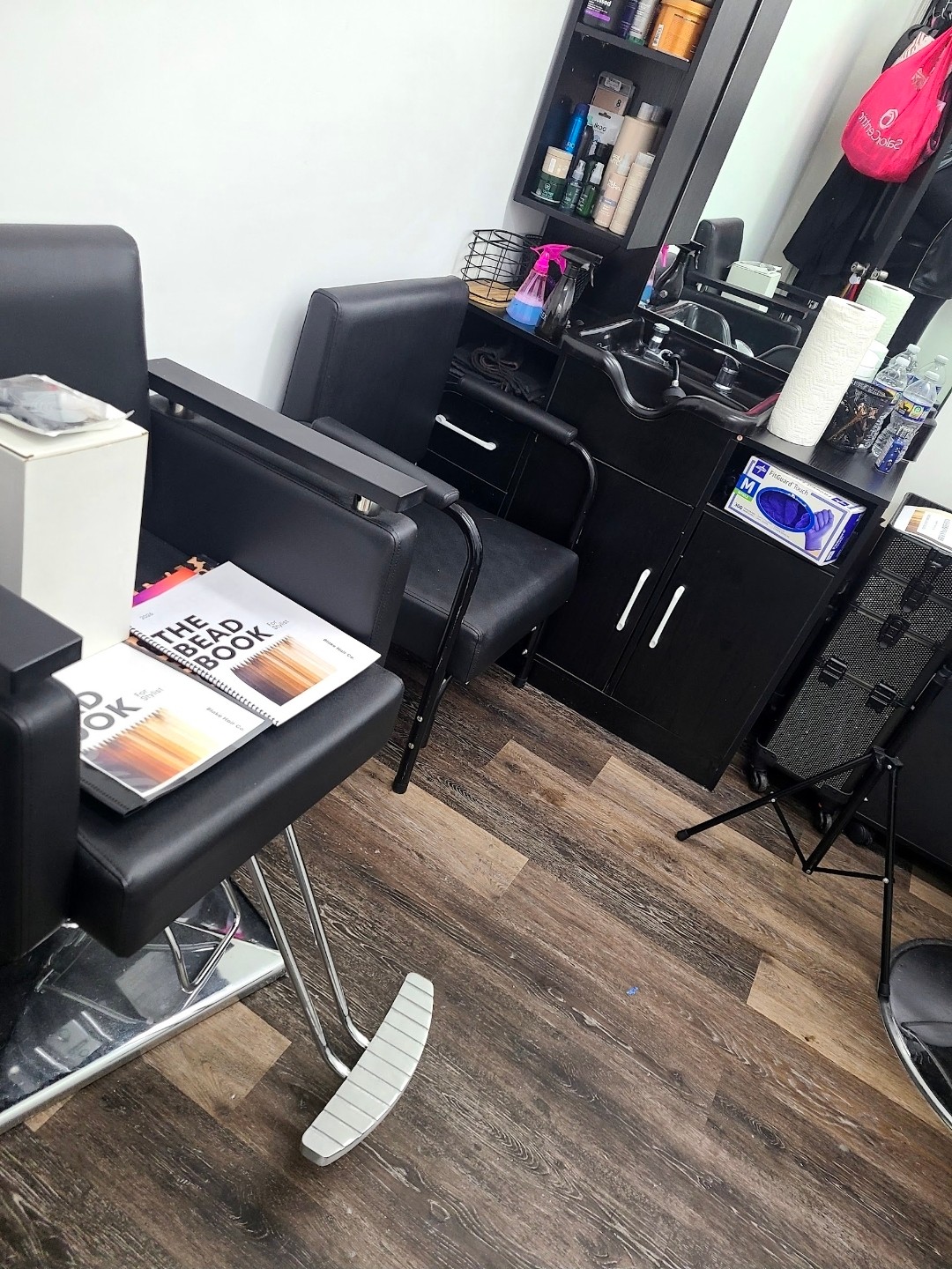 Salon station with chairs, mirror, and hair products, including a pink bag hanging on the wall, and a wooden floor.
