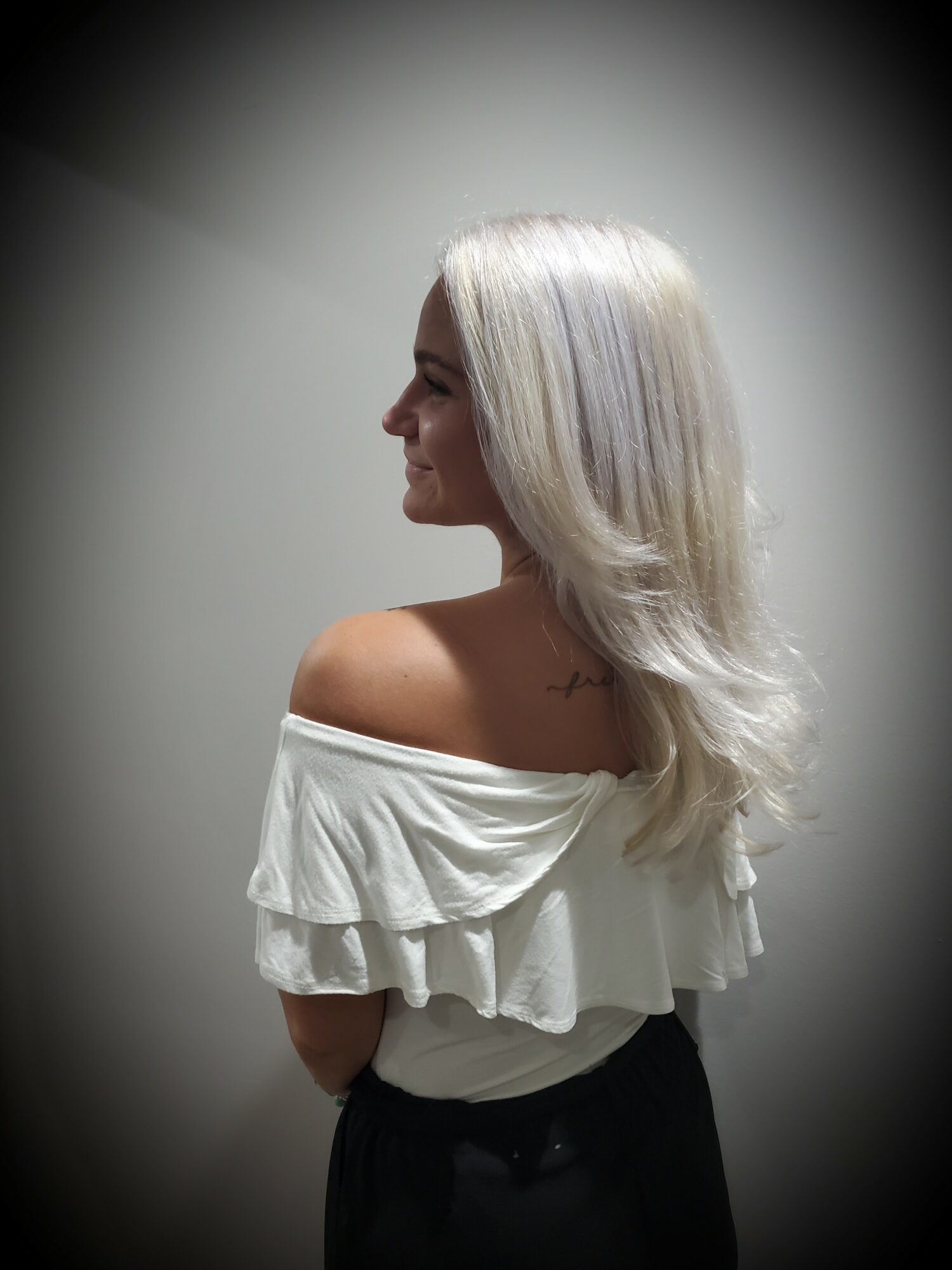 Side profile of a woman with long, wavy, platinum blonde hair, wearing an off-the-shoulder white top, against a plain background.