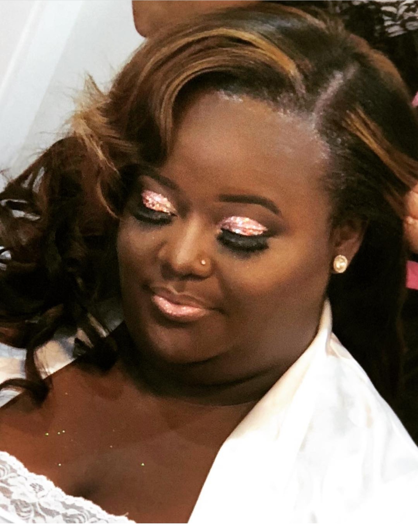 Woman with styled hair, makeup, and earrings, looking downward with a calm expression.