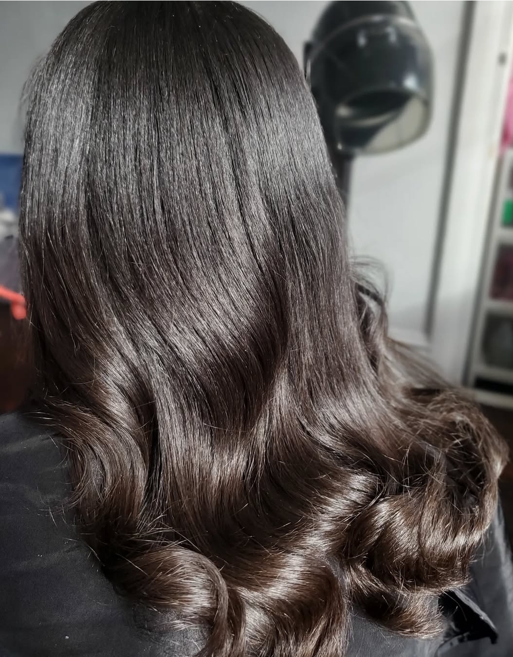 Person with long, shiny, wavy dark hair, viewed from the back in a salon setting.