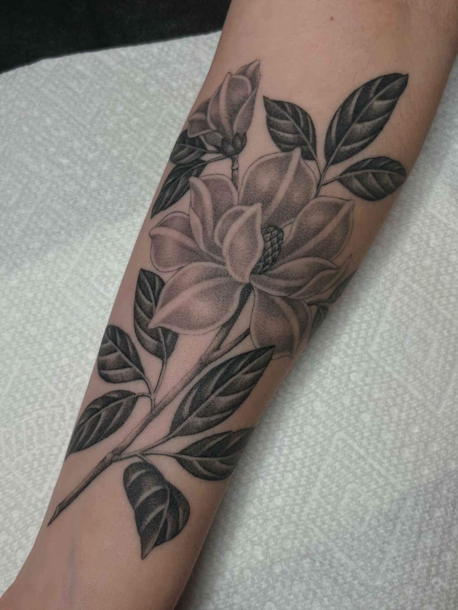 Black and gray floral tattoo with large flowers and leaves on person's forearm.