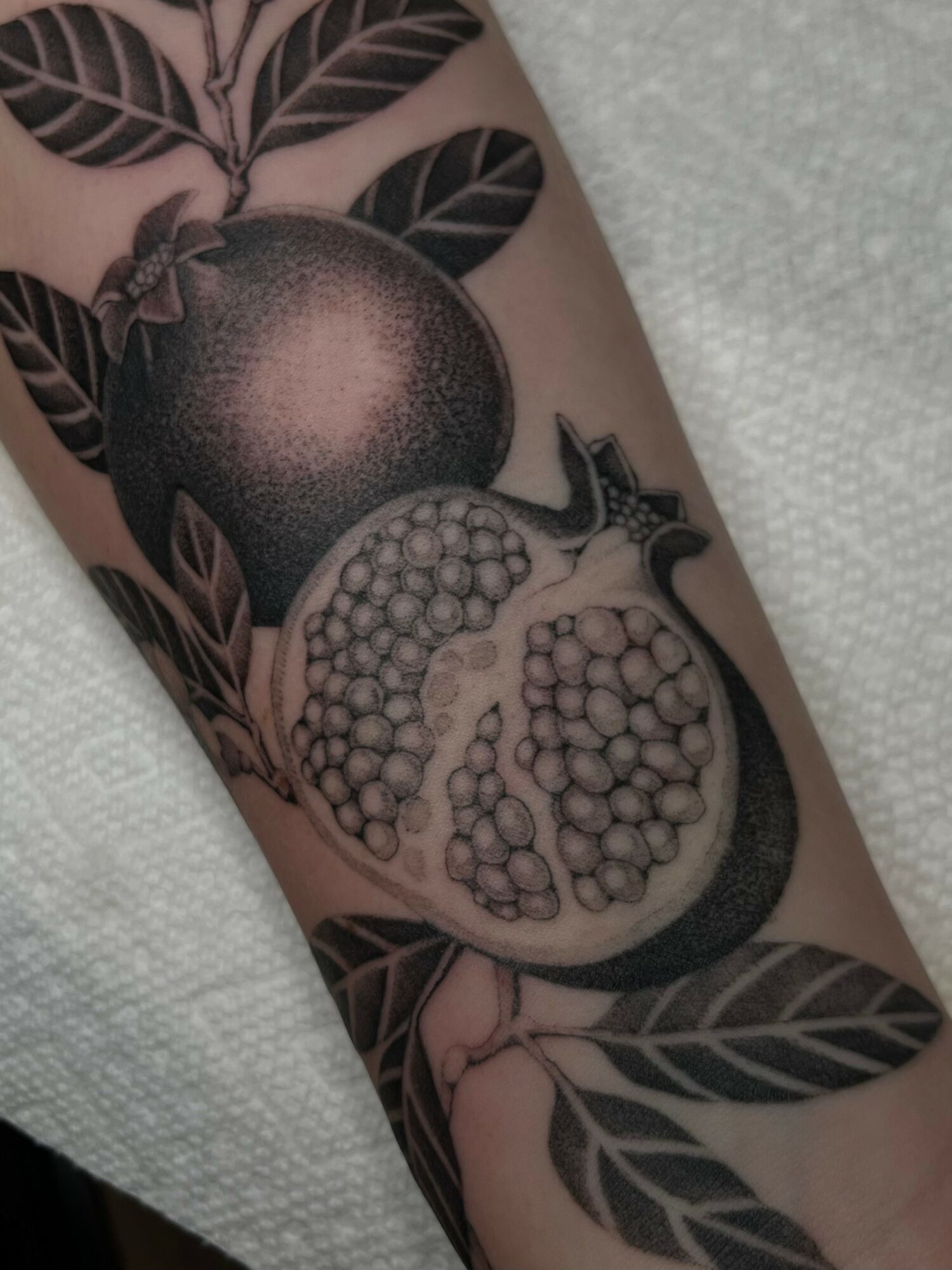 Pomegranate tattoo with leaves on skin, partially open showing seeds inside, in black and gray ink.